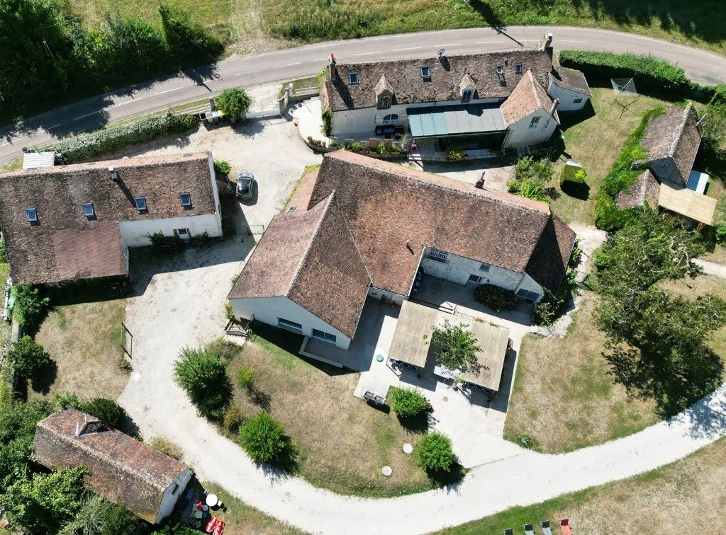 Photo aérienne de plusieurs maisons rurales avec toits en tuiles, jardins et zones de stationnement, entourées de verdure et d'une route sinueuse.