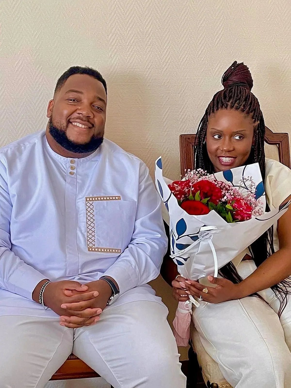 Un homme et une femme assis côte à côte, la femme tient un bouquet de fleurs rouges et roses, tous deux souriants. La scène semble être prise dans un intérieur.