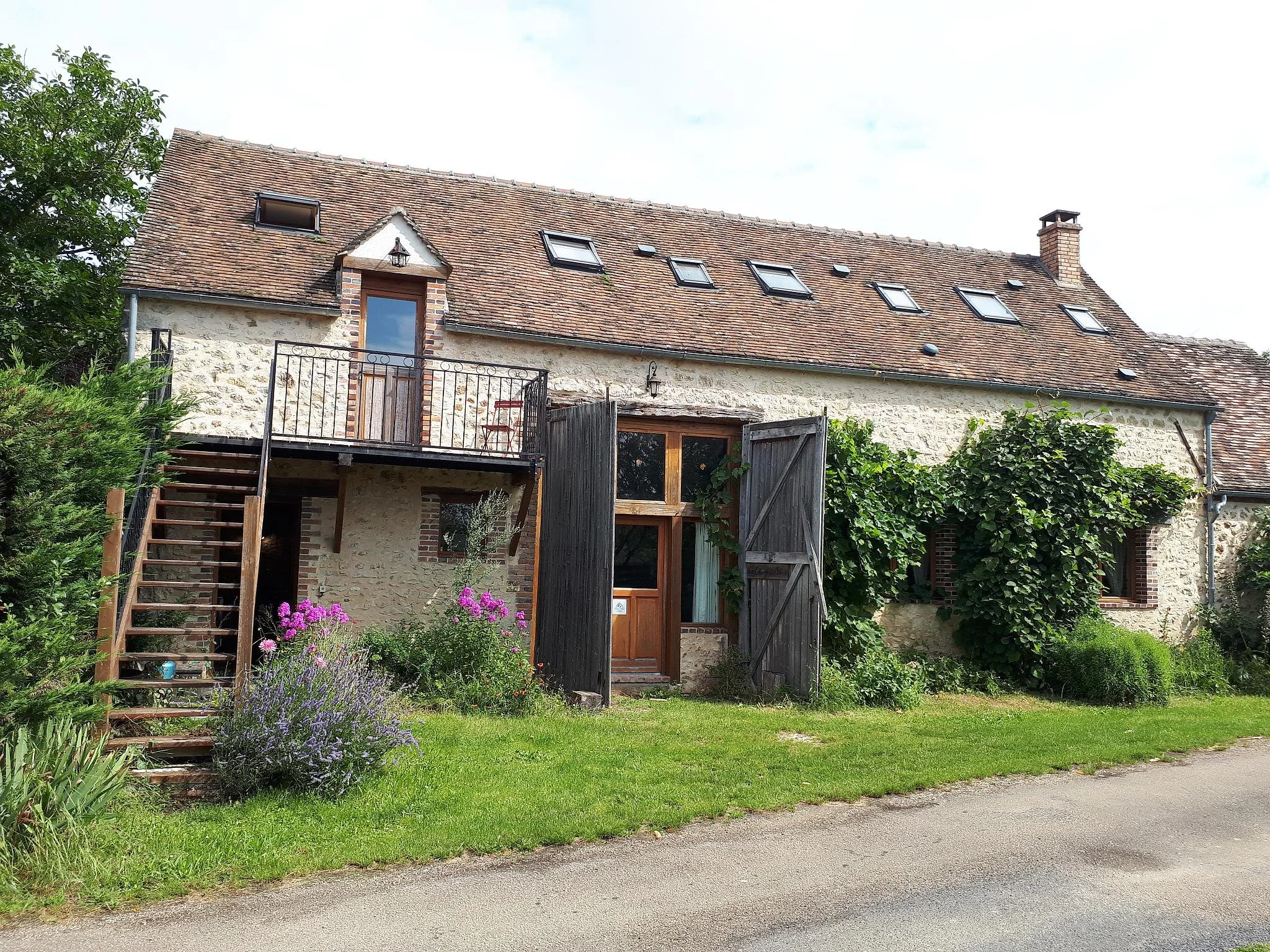 Maison en pierre avec une toiture en tuiles, volets en bois, escalier en bois, et végétation luxuriante autour, dont des plantes grimpantes et des fleurs colorées.