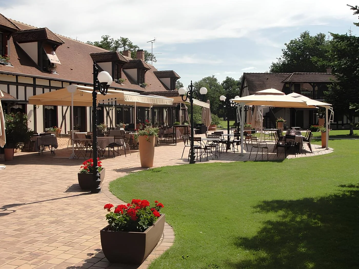 Terrasse extérieure d'un restaurant ou café avec des tables, chaises, parasols, lampadaires et fleurs décoratives, entourée d'un bâtiment de style traditionnel avec jardins bien entretenus.