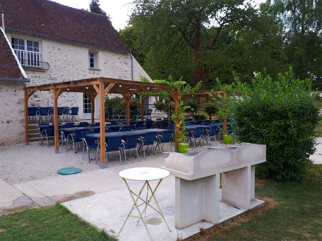 Une terrasse extérieure avec une pergola en bois, des tables et des chaises bleues, un mur de maison en pierre, et un espace vert avec des arbres et des buissons.
