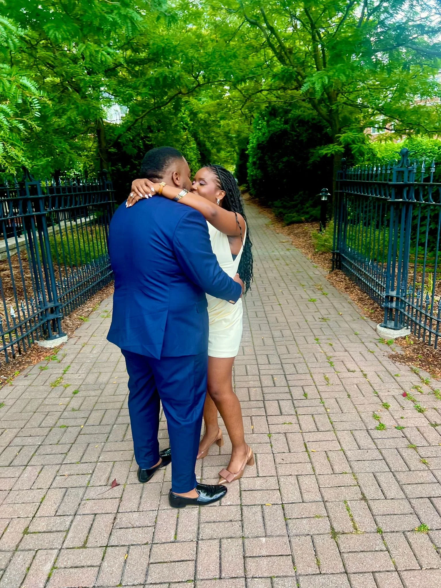 Un couple s'embrassant dans un parc verdoyant, la femme en robe blanche et talons beiges, l'homme en costume bleu, sous un arbre.