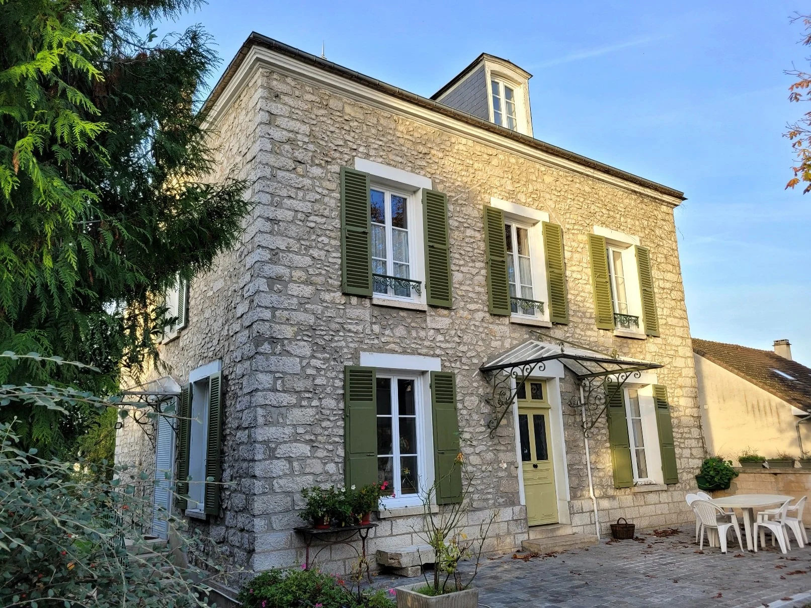 Une maison en pierre avec des volets verts, une porte d'entrée beige, une cour avec une table en plastique blanche et des chaises, des plantes en pot, dans un cadre en plein air ensoleillé.