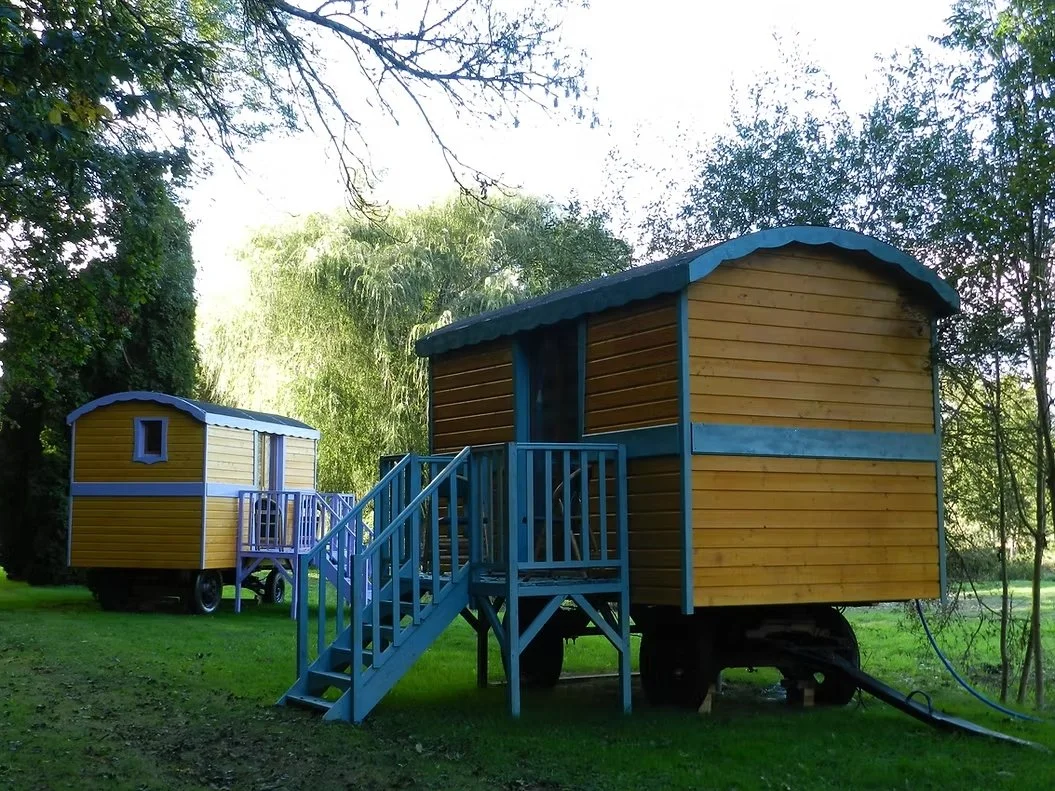 Deux petites maisons en bois colorées sur des roues, avec des escaliers en bois, situées dans un jardin verdoyant, entouré d'arbres.