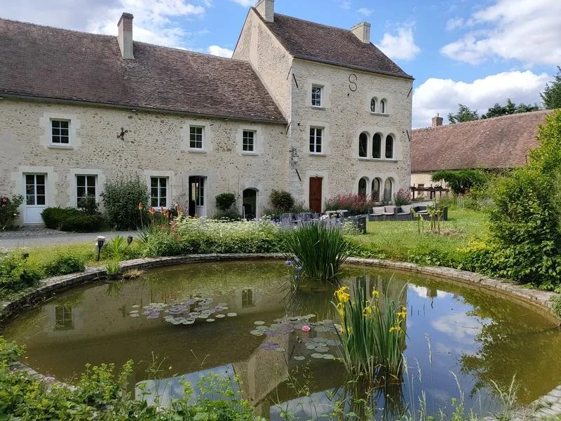 Une maison ancienne en pierre avec plusieurs fenêtres, entourée d'un jardin avec un petit étang et des plantes, sous un ciel partiellement nuageux.