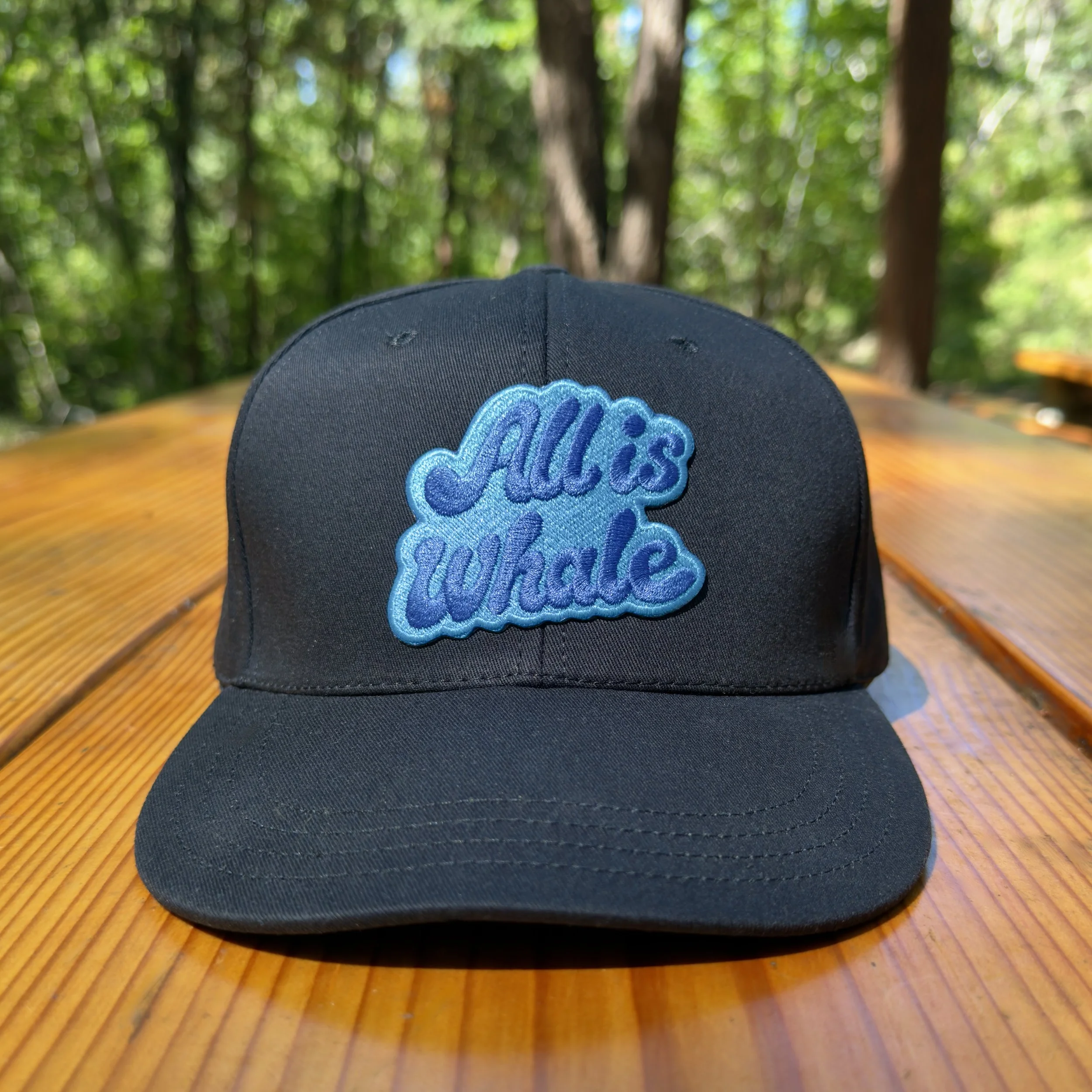 Image of a black baseball cap with an "All is whale" message sitting atop a picnic table