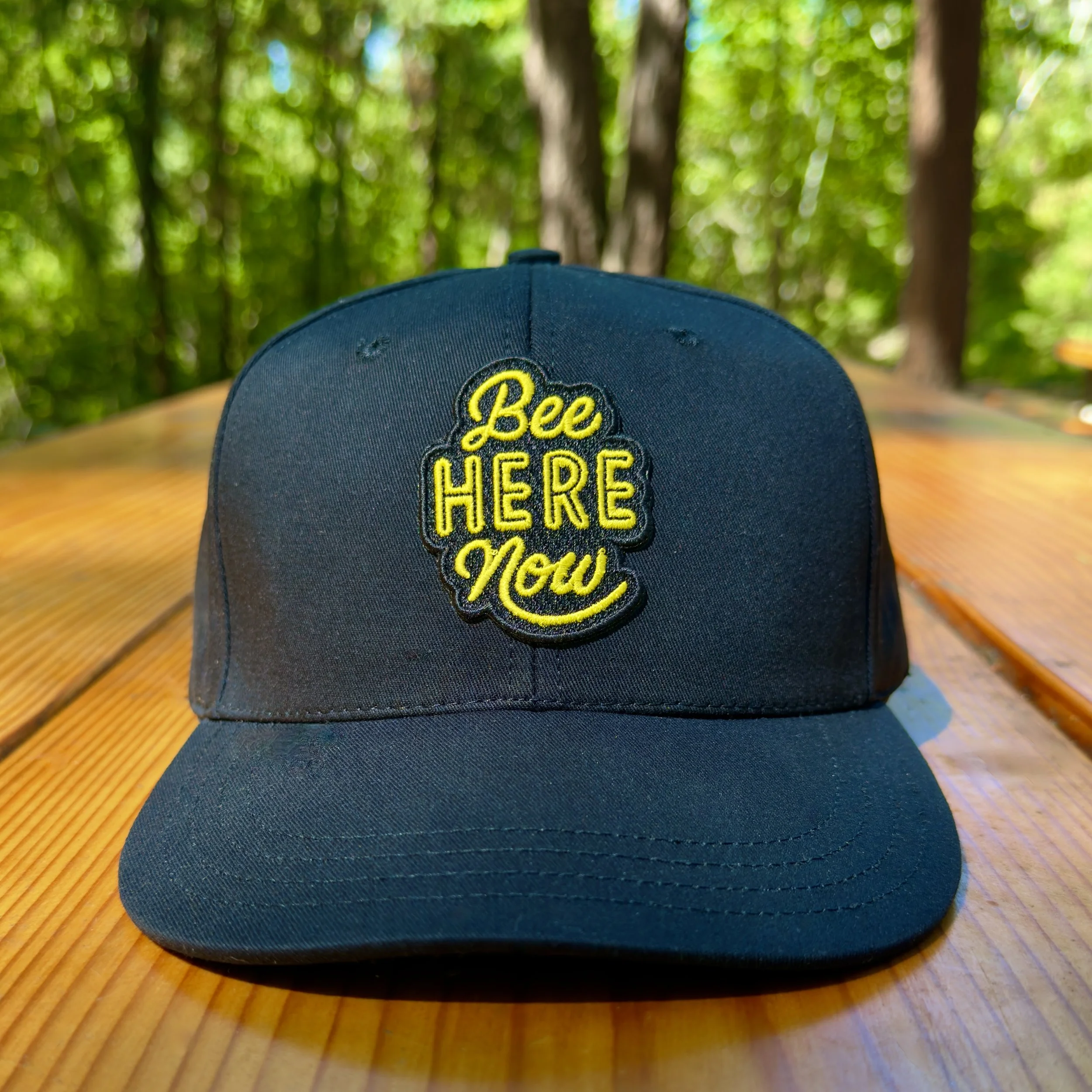 Image of a black baseball cap with a "Bee here now" message sitting atop a picnic table