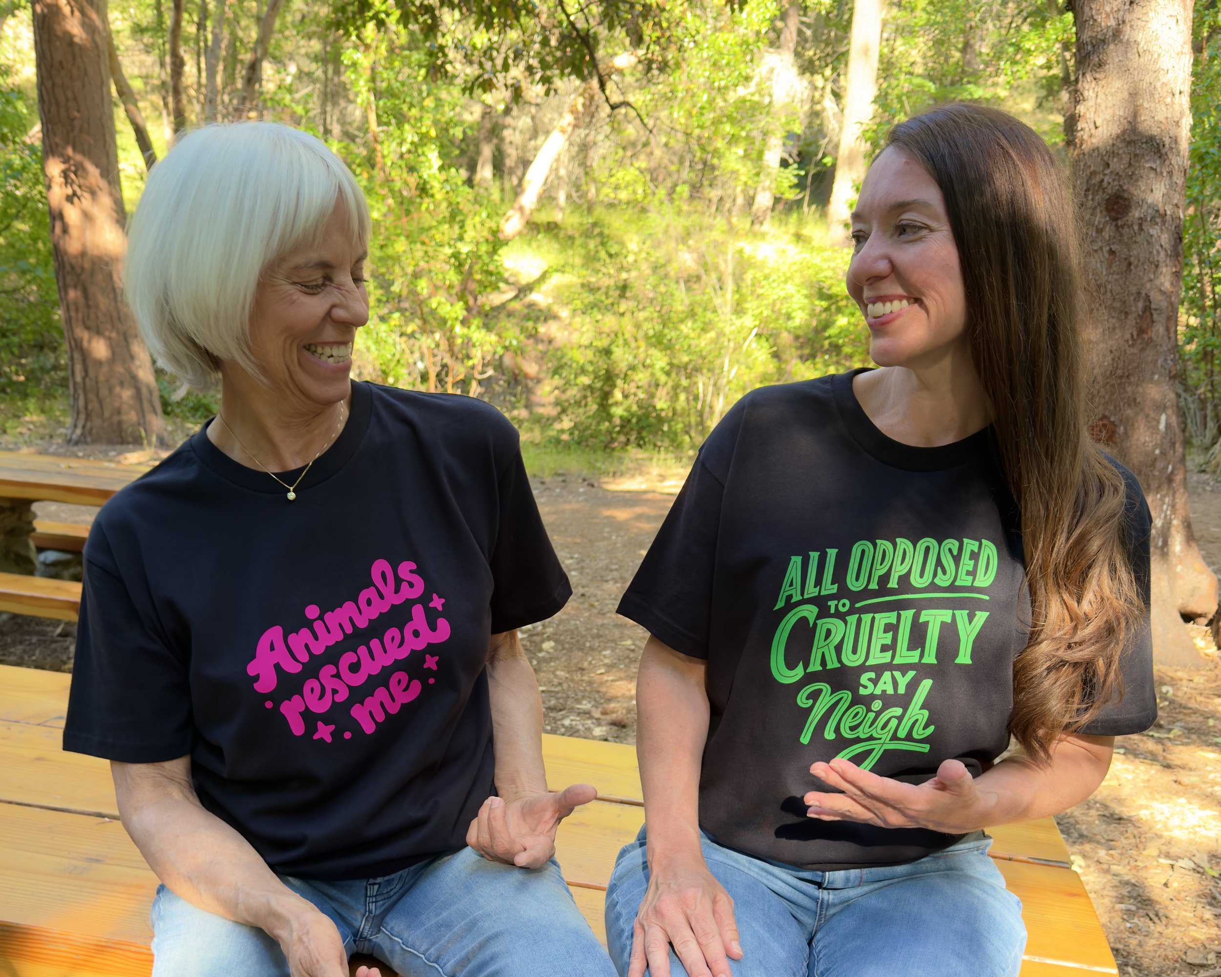 Two women sit on a picnic table smiling at each other. They are wearing black t-shirts. The message on one shirt is: Animals rescued me. The message on the other shirt is: All opposed to cruelty say neigh.