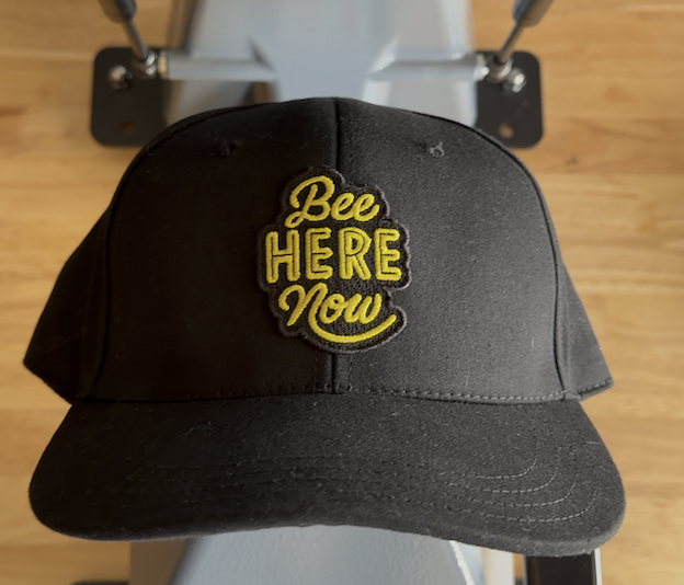 A hat press has a black ball cap on it with a patch that reads: Bee here now