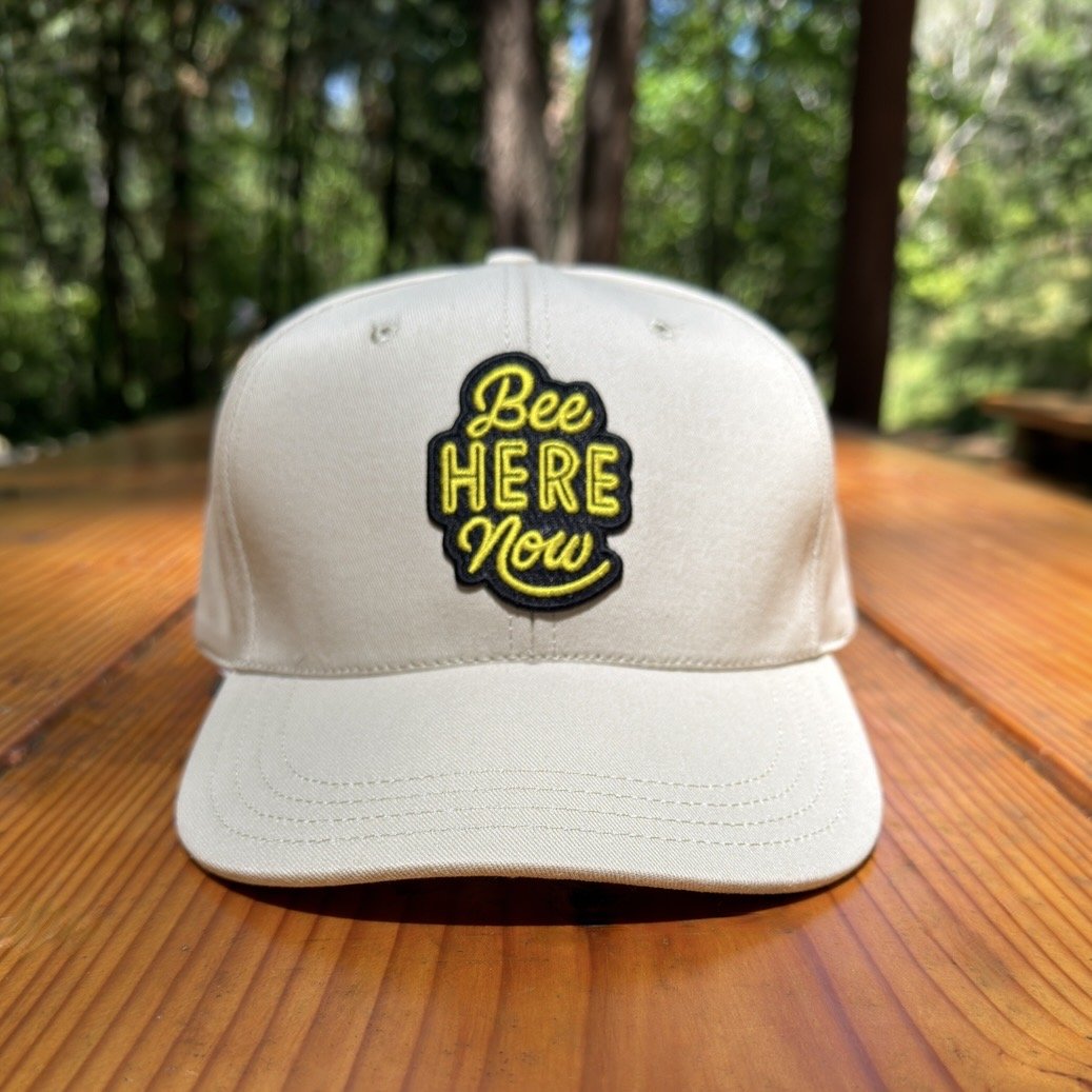 Image of an off-white baseball cap with an "Bee here now" message sitting atop a picnic table