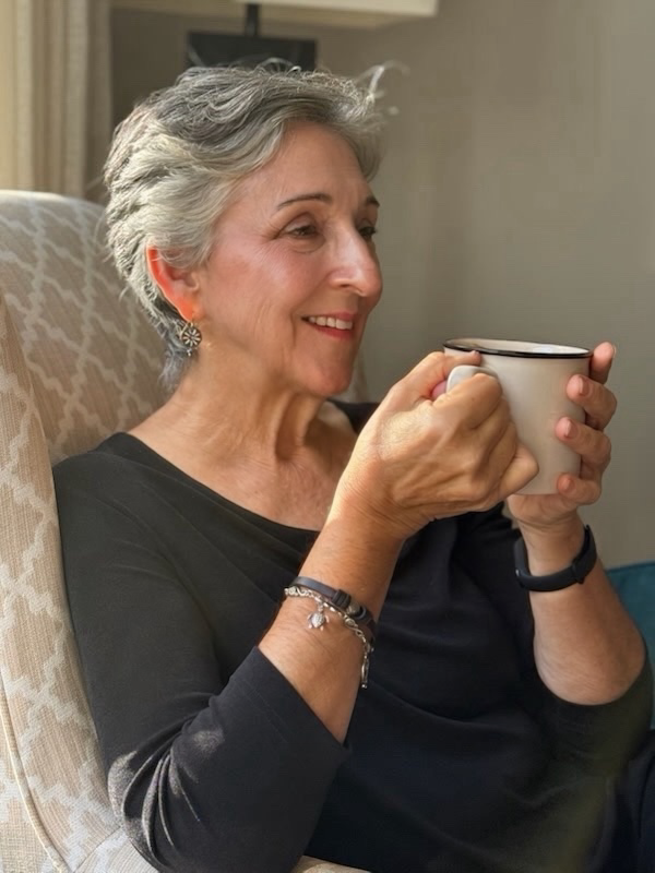 Older Woman sitting in a chair with a slight smile and reflective gaze holding a coffee cup