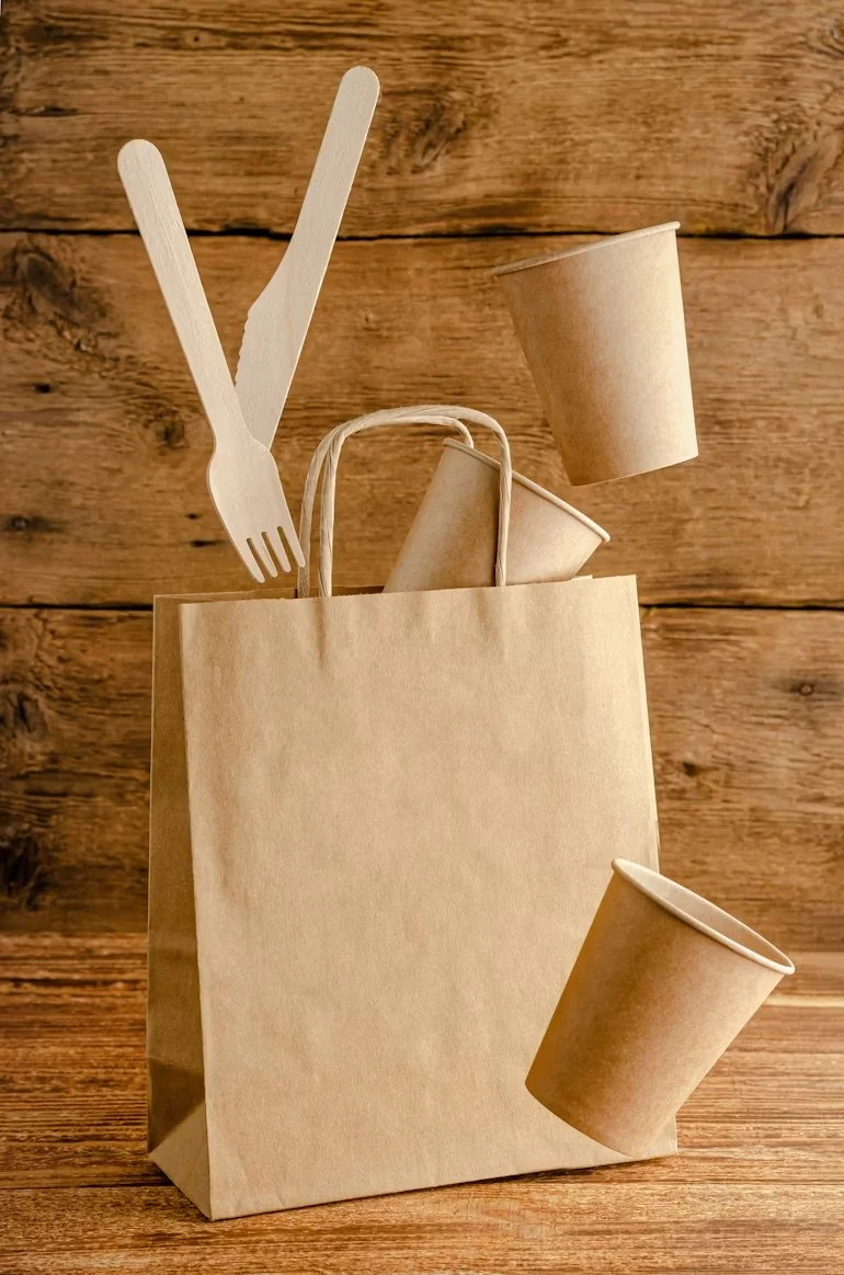 A brown paper bag with wooden utensils and three paper cups on a wooden background.