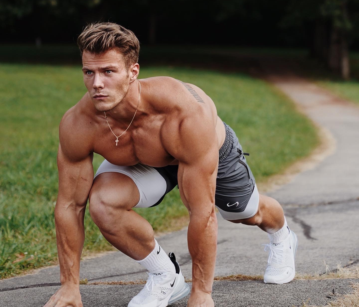 A shirtless man with light skin, muscular build, and short light brown hair is outside on a paved path in a crouched position, preparing to run or jump. He wears white and gray athletic shorts, white sneakers, white socks, and a necklace with a cross pendant. The background features grass and trees.
