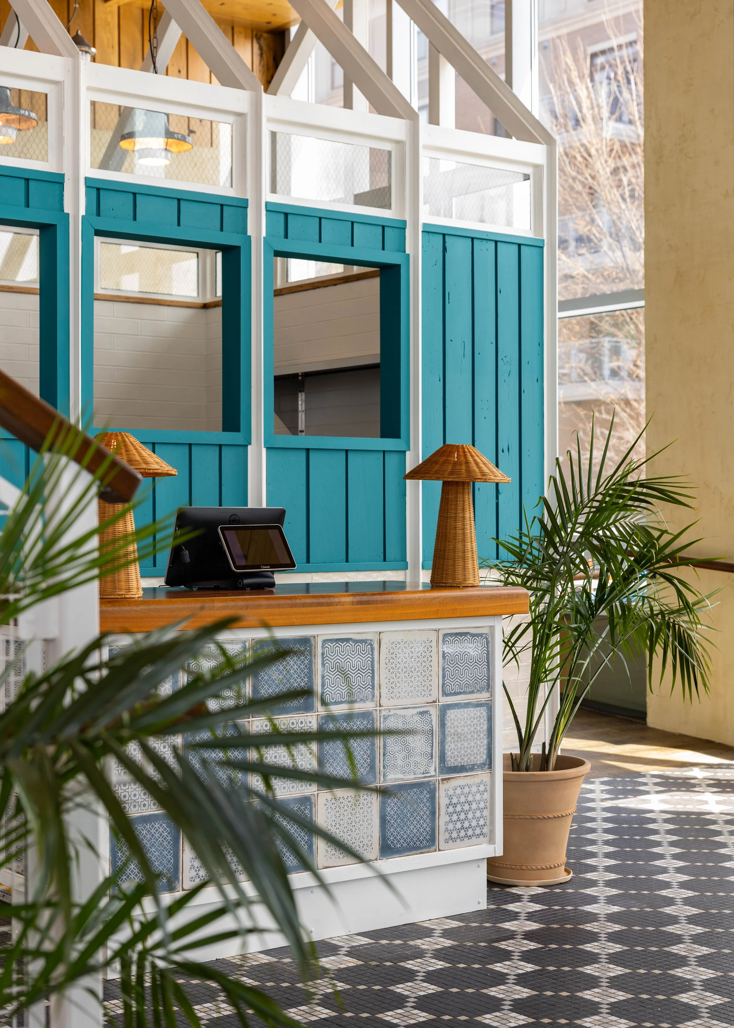 Interior of a modern cafe or restaurant with a checkered black and white tile floor, potted plants, a counter with a tiled front, and a decorative blue wooden partition with square cutouts and windows.