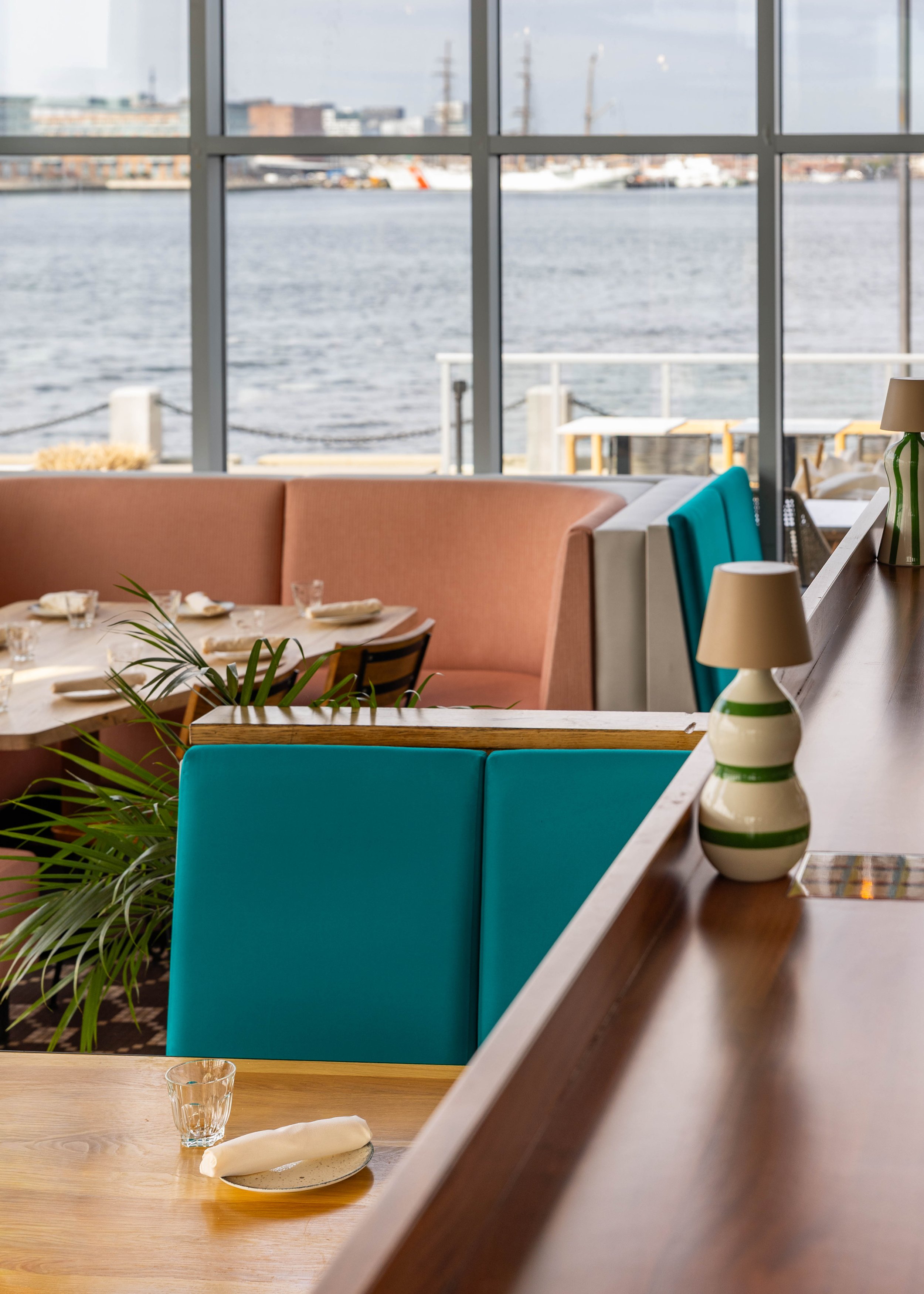 Interior of a restaurant with a view of the water outside large windows, featuring a table set with glasses, napkins, and plates, colorful chairs, and decorative lamps on the counter.