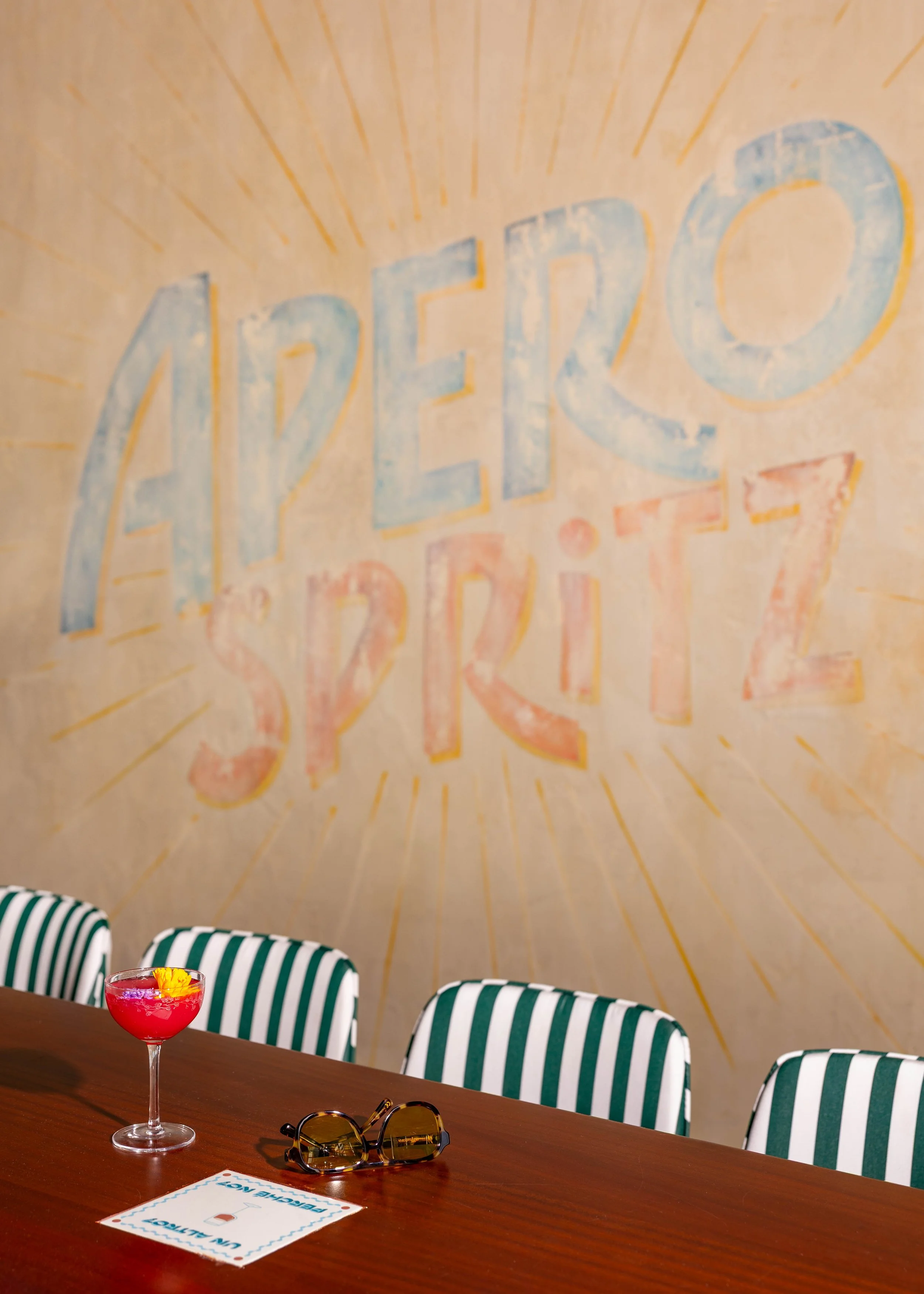 A bar or restaurant with a wall art that reads 'ARETO SPIRITS' in pastel colors, a cocktail with a flower on top, sunglasses, and a paper sign on a wooden table, with striped chairs in the background.