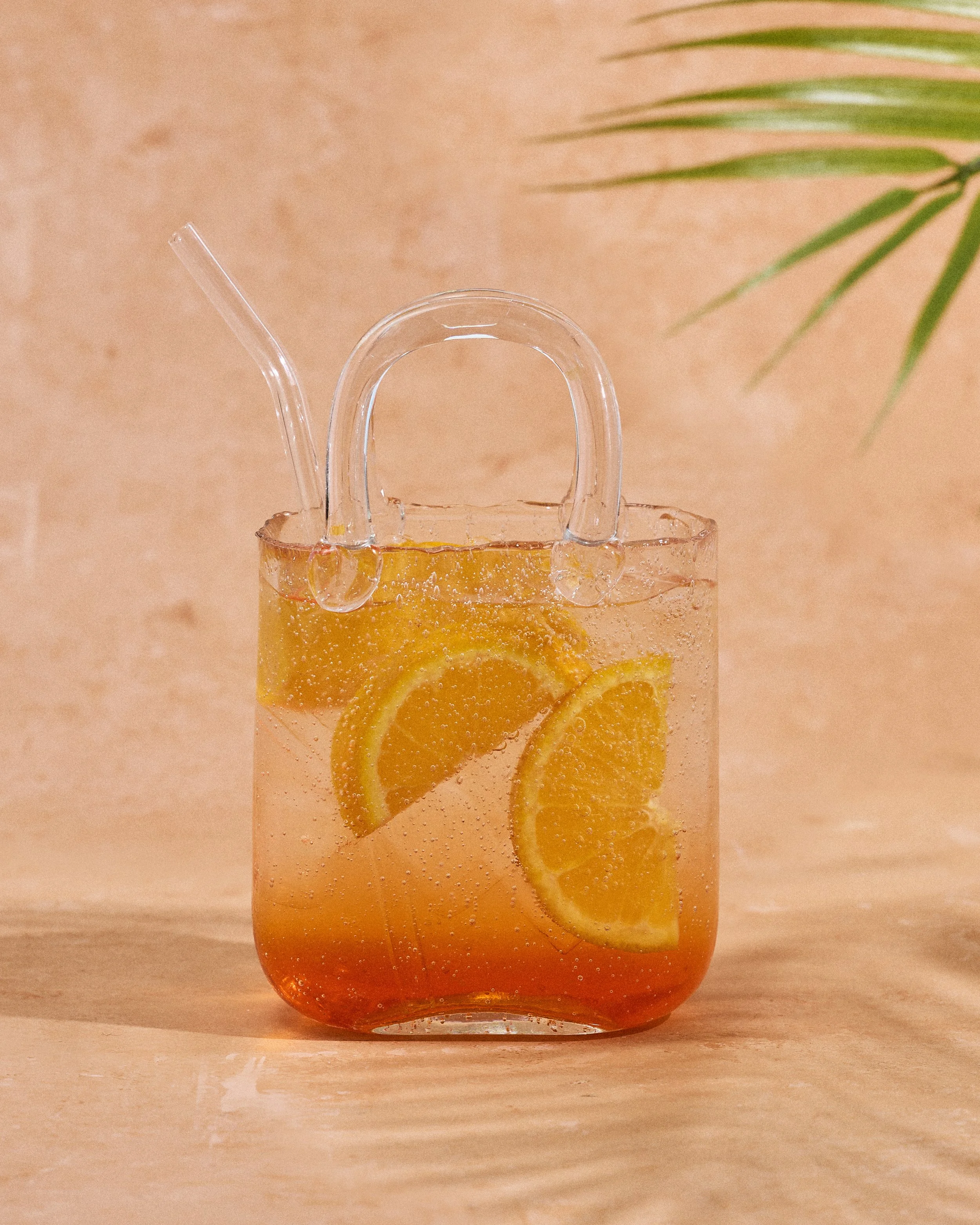 Glass pitcher filled with orange slices and a sparkling orange drink, with a clear straw, on a light wooden surface, with a peach-colored background and a green plant leaf in the top right corner.