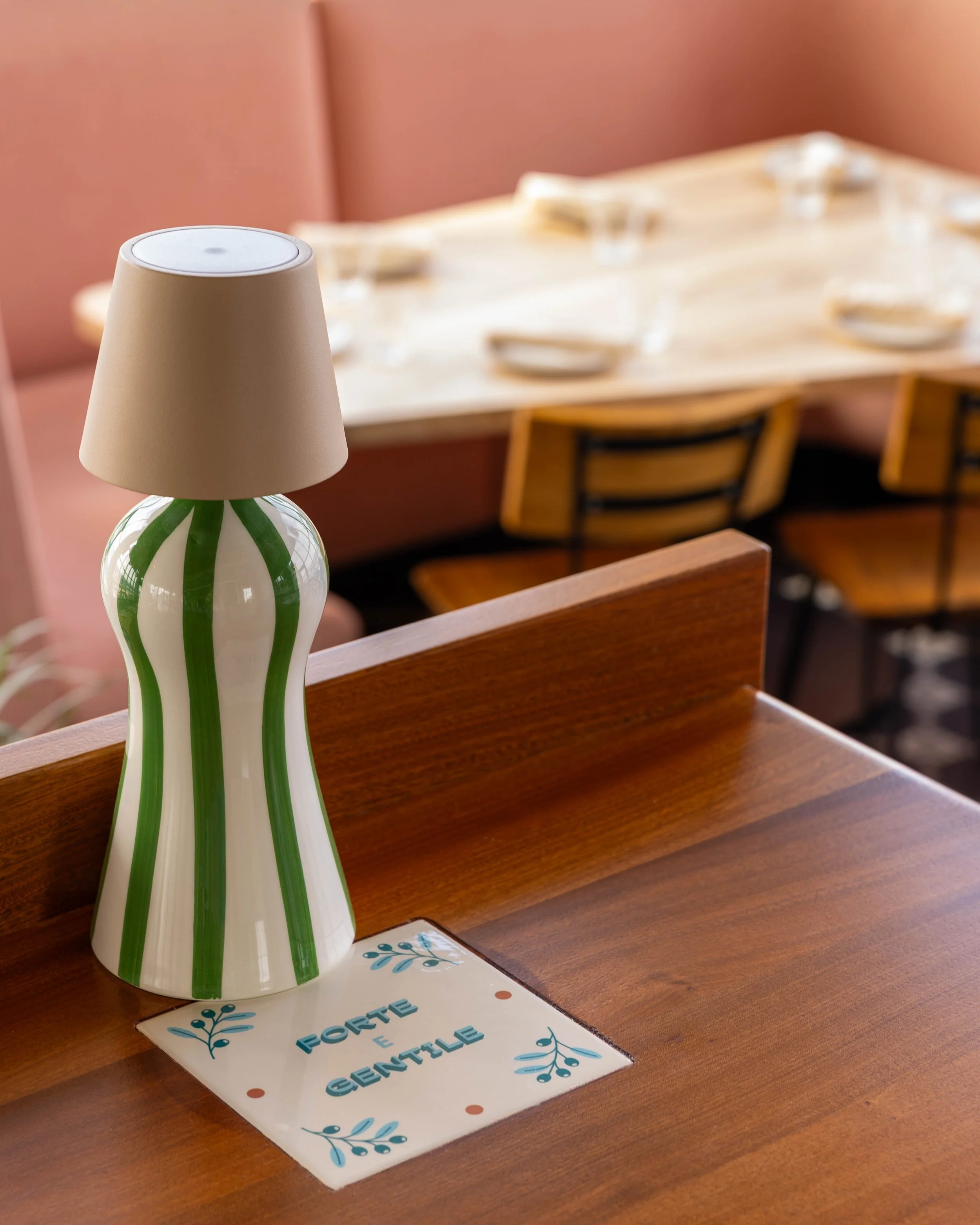 A decorative lamp with a beige lampshade and a ceramic base with green and white wavy stripes on a wooden table. A sign on the table reads 'Forte e Gentile' with blue and red accents, and the background shows a dining area with a table set with plate