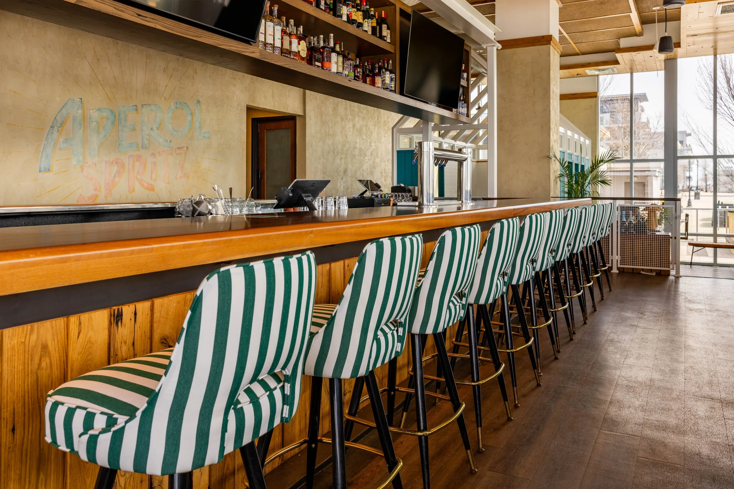Empty bar with striped green and white chairs, bottles on shelves, and large windows letting in natural light.