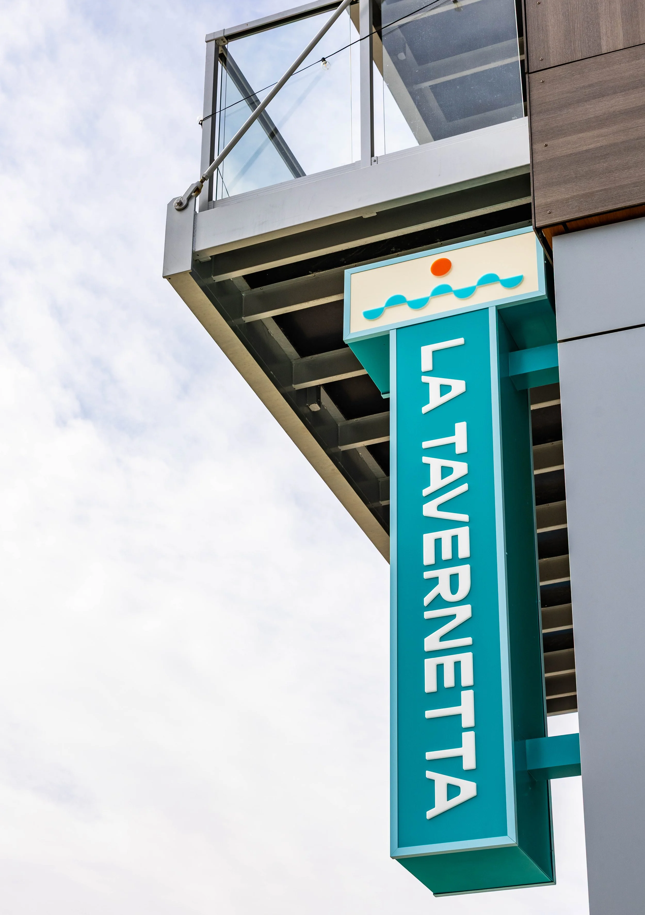 Photo of a sign for a swimming pool called 'La Taverneeta' with a symbol of water waves and a sun above it, attached to a modern building.