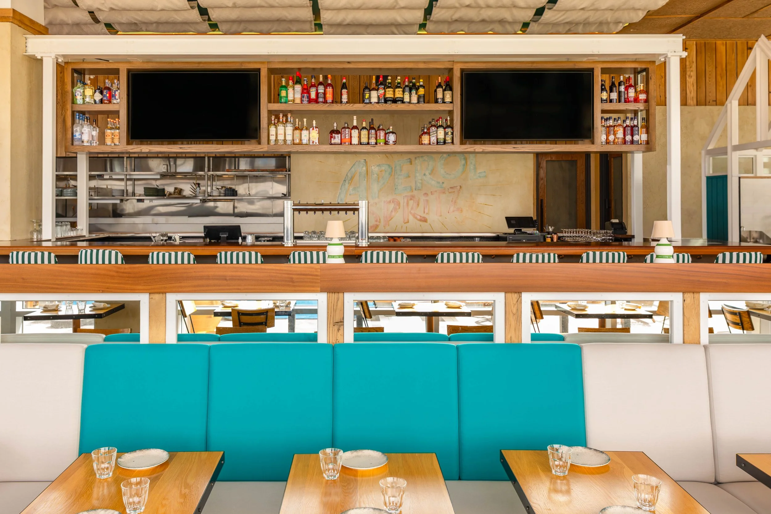 Empty restaurant with tables set for dining, a teal booth in the foreground, and a bar in the background with shelves of various liquor bottles and two large black screens.