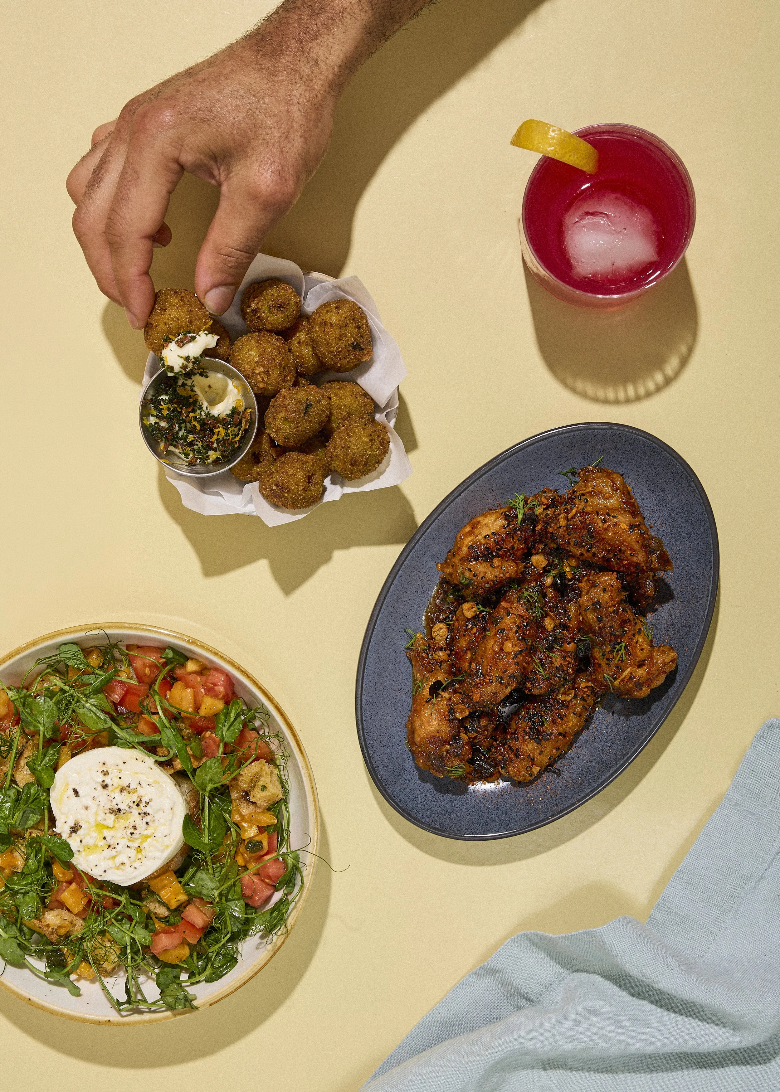 A flat lay of an assortment of food and drink on a yellow table, including a plate of fried croquettes with sauce, a bowl of salad with a poached egg, a plate of spicy glazed chicken wings, and a pink cocktail with a lemon wedge.