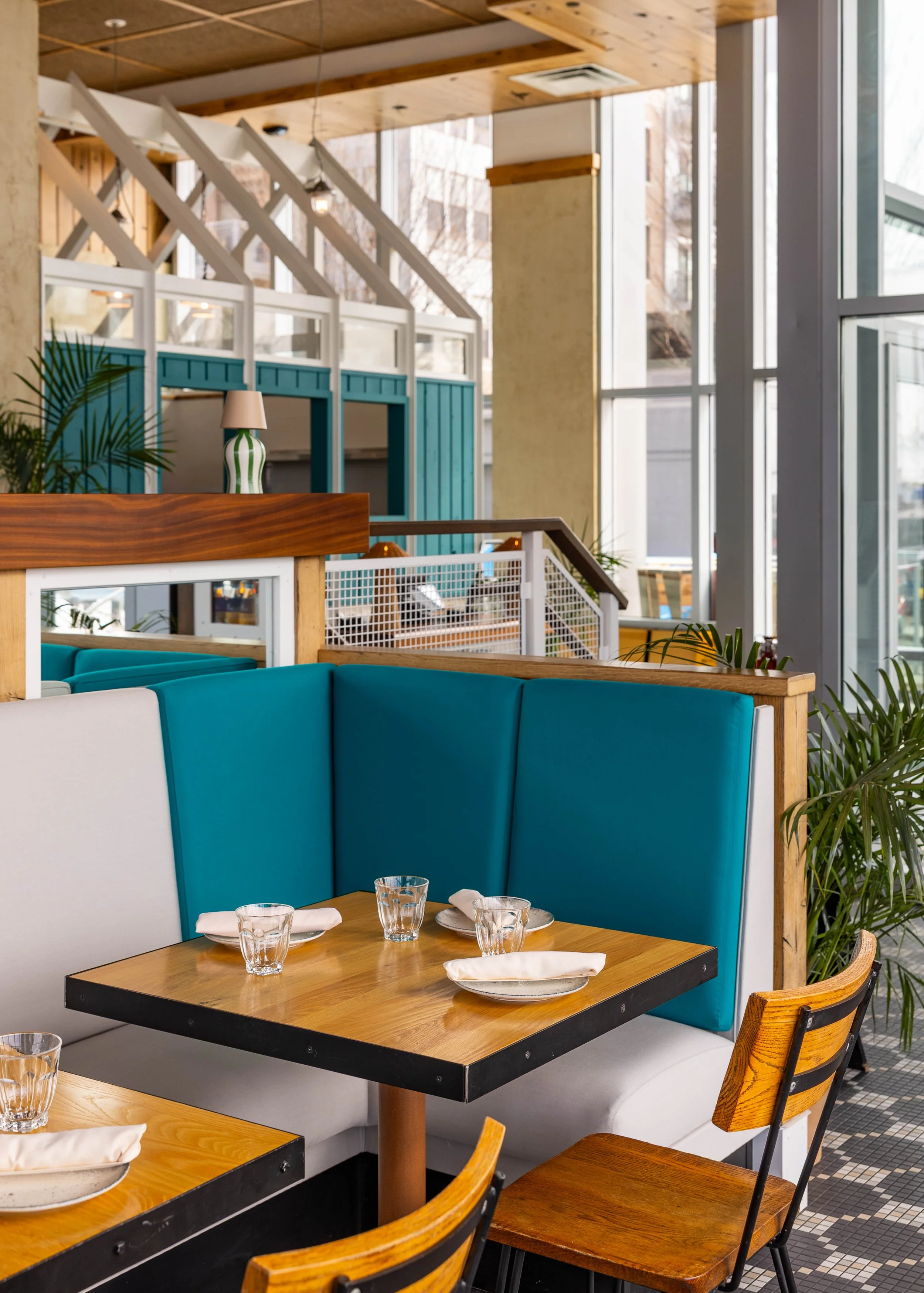 Interior of a modern restaurant with teal booth seating and wooden tables set with napkins, glasses, and plates, large windows, and decorative plants.