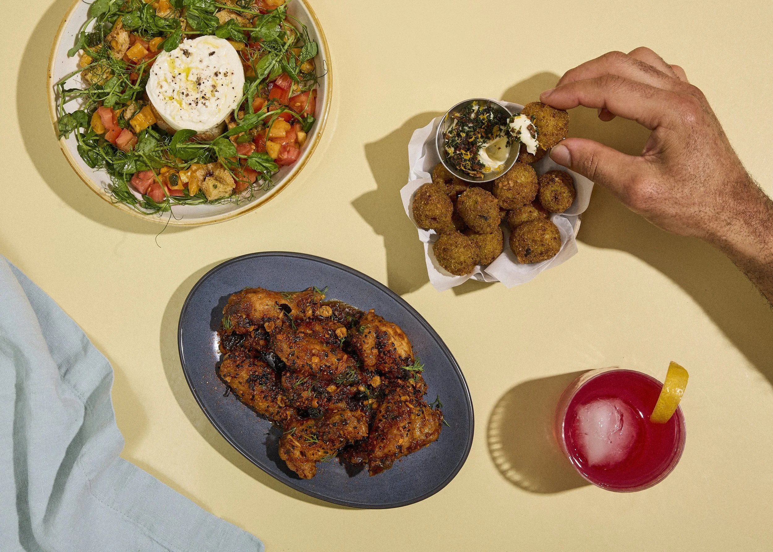 Overhead view of a table with a salad, fried food, chicken wings, and a pink cocktail with lemon on a yellow table