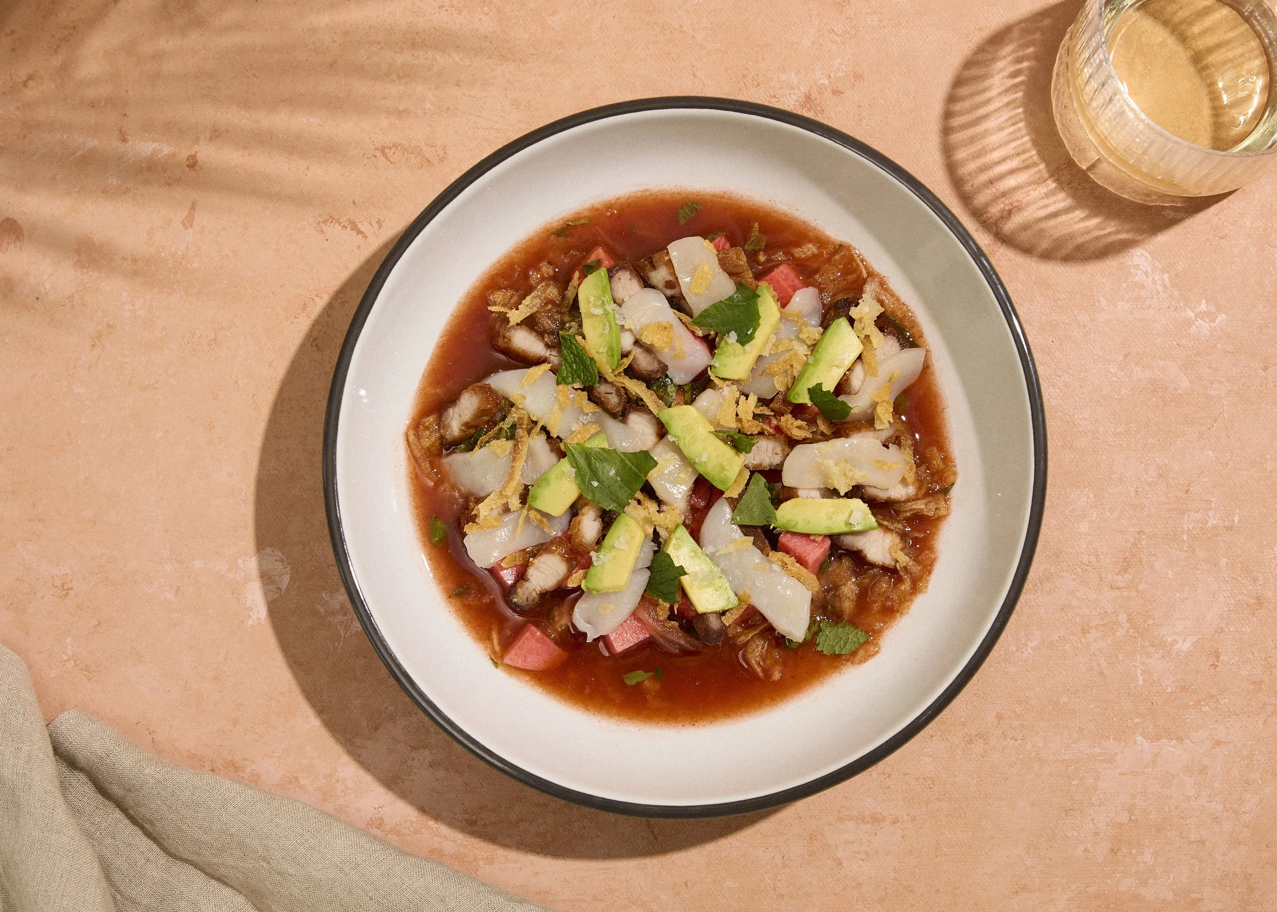 A bowl of Mexican cochinita pibil with shredded pork, pickled onions, tomato, avocado, and herbs on a peach-colored surface with a glass of pale yellow beverage.
