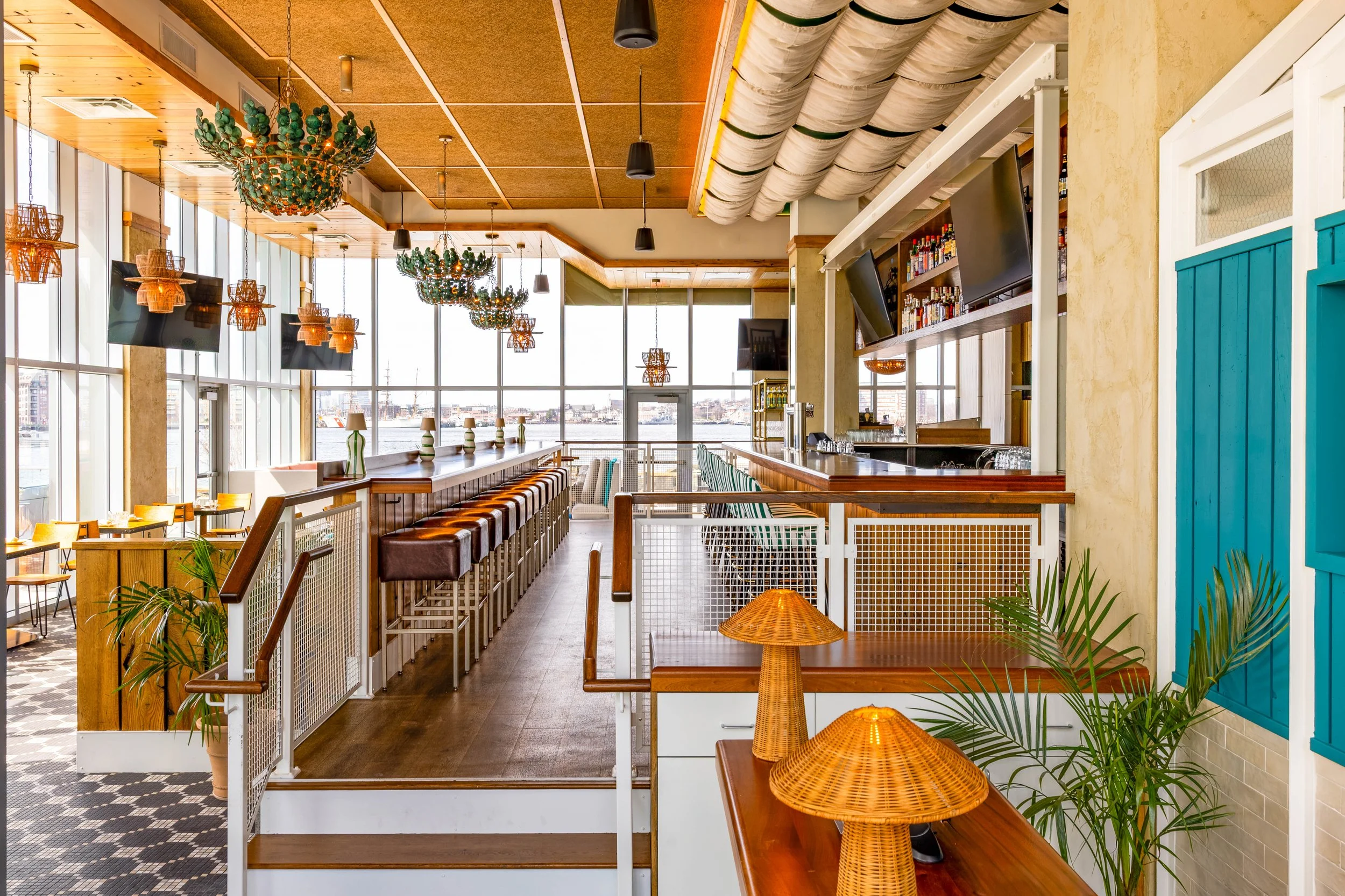 Interior of a modern restaurant or bar with large windows, wooden accents, hanging lights, and seating along a bar counter. Indoor plants and decorative lamps are also visible.