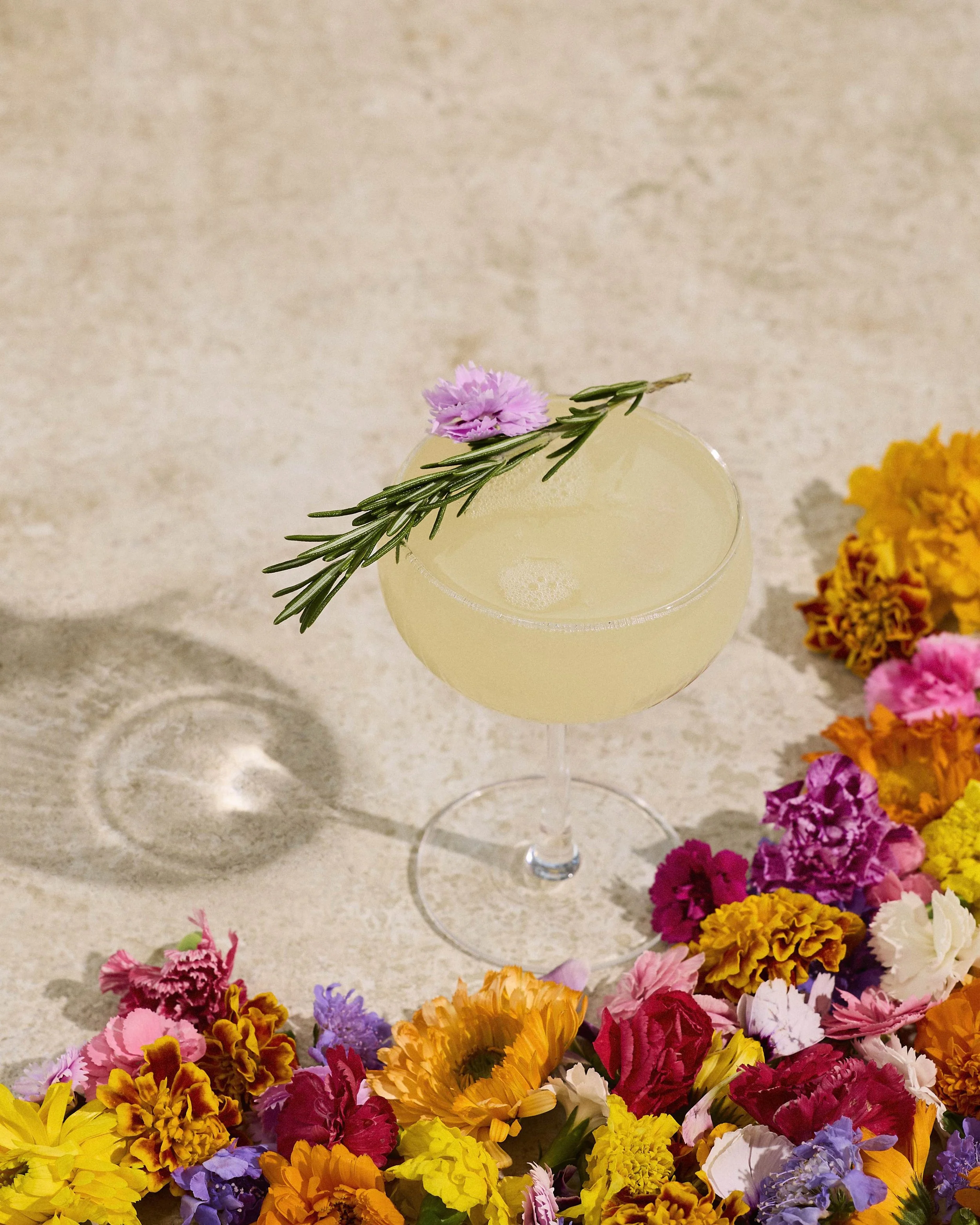 A pale yellow cocktail garnished with a sprig of rosemary and a pink flower, surrounded by colorful flowers on a light-colored surface.