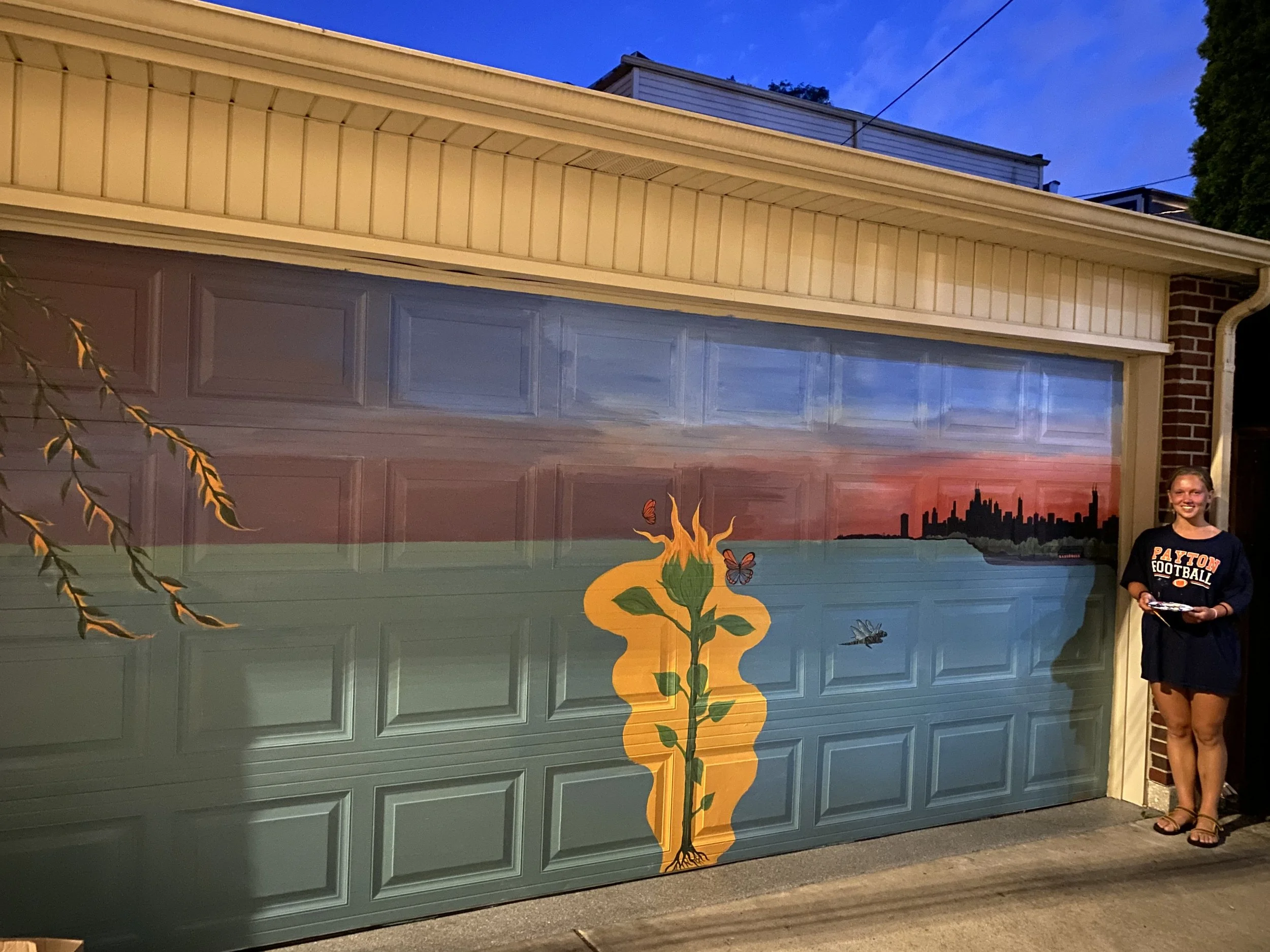 A painted mural on a garage door depicting a sunset skyline in the distance, with a large sunflower in the center surrounded by butterflies and insects. A woman in a black sweatshirt and shorts stands to the right, smiling and holding a paper.