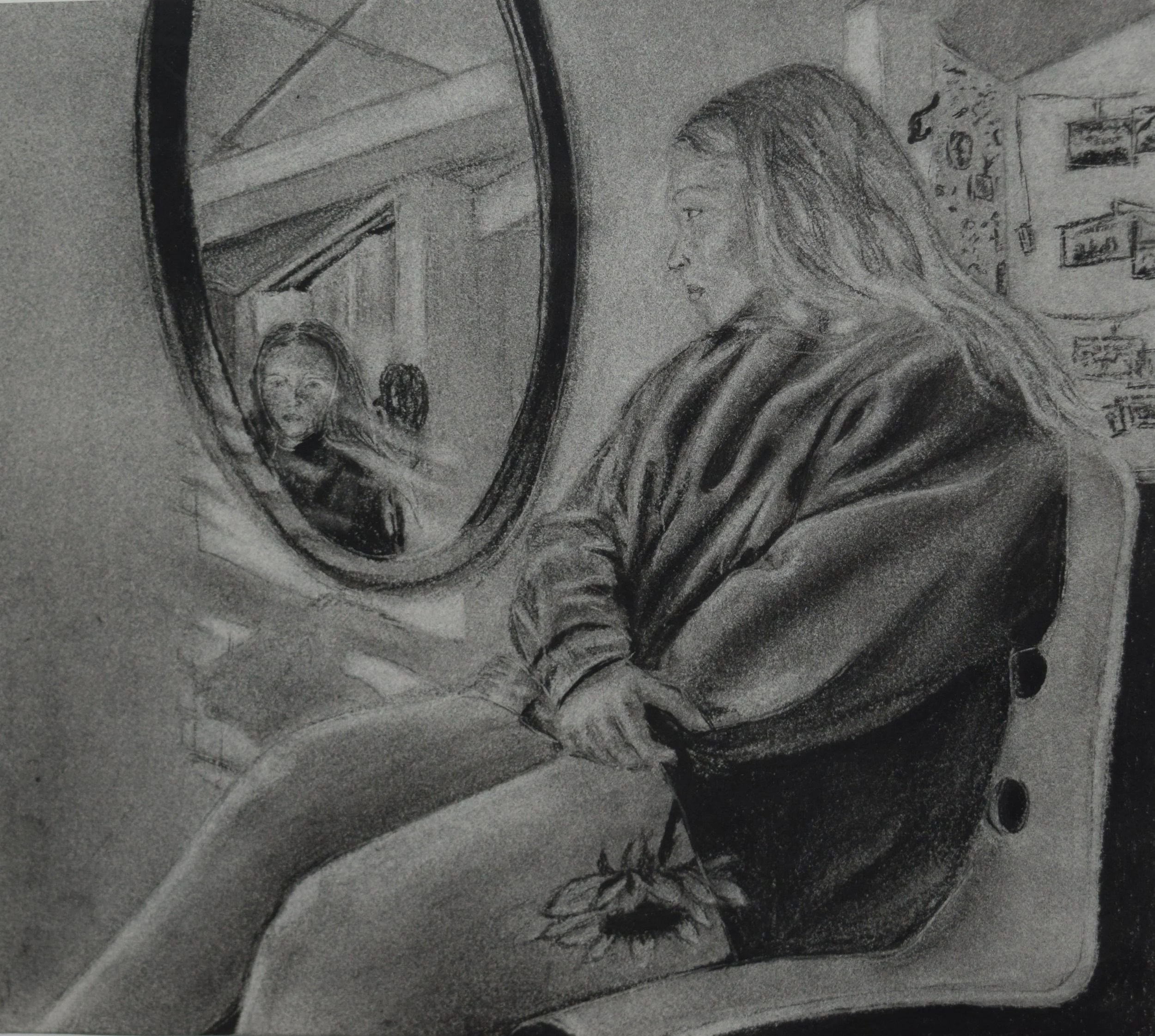 A woman with long hair looking into a mirror, holding a dying sunflower to her thigh, seated on a chair in a room with photos and artwork on the walls.