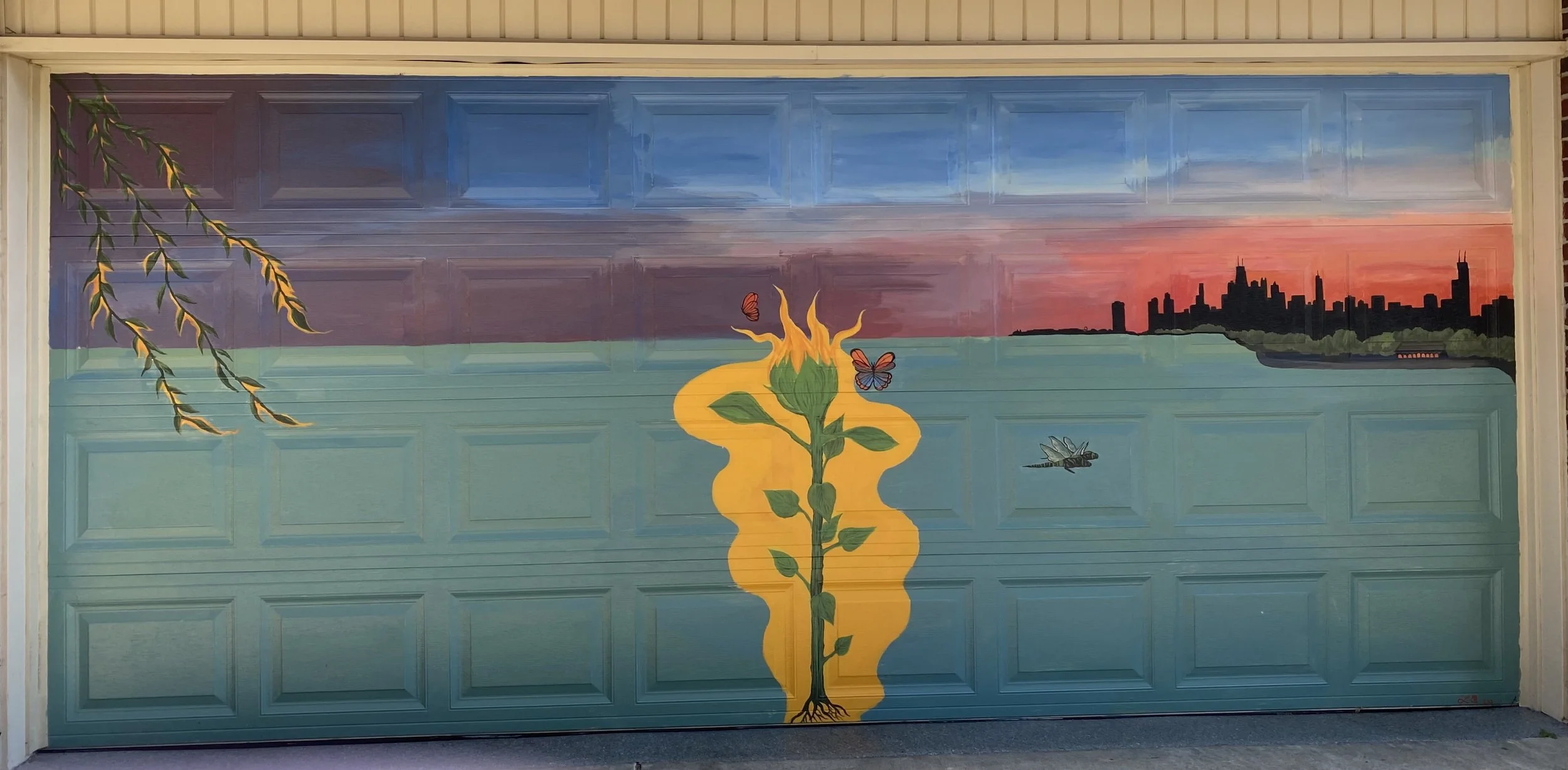A mural painted on a garage door depicting a sunset over a city skyline with a body of water, a sunflower in the center with butterflies, and a dragonfly nearby.