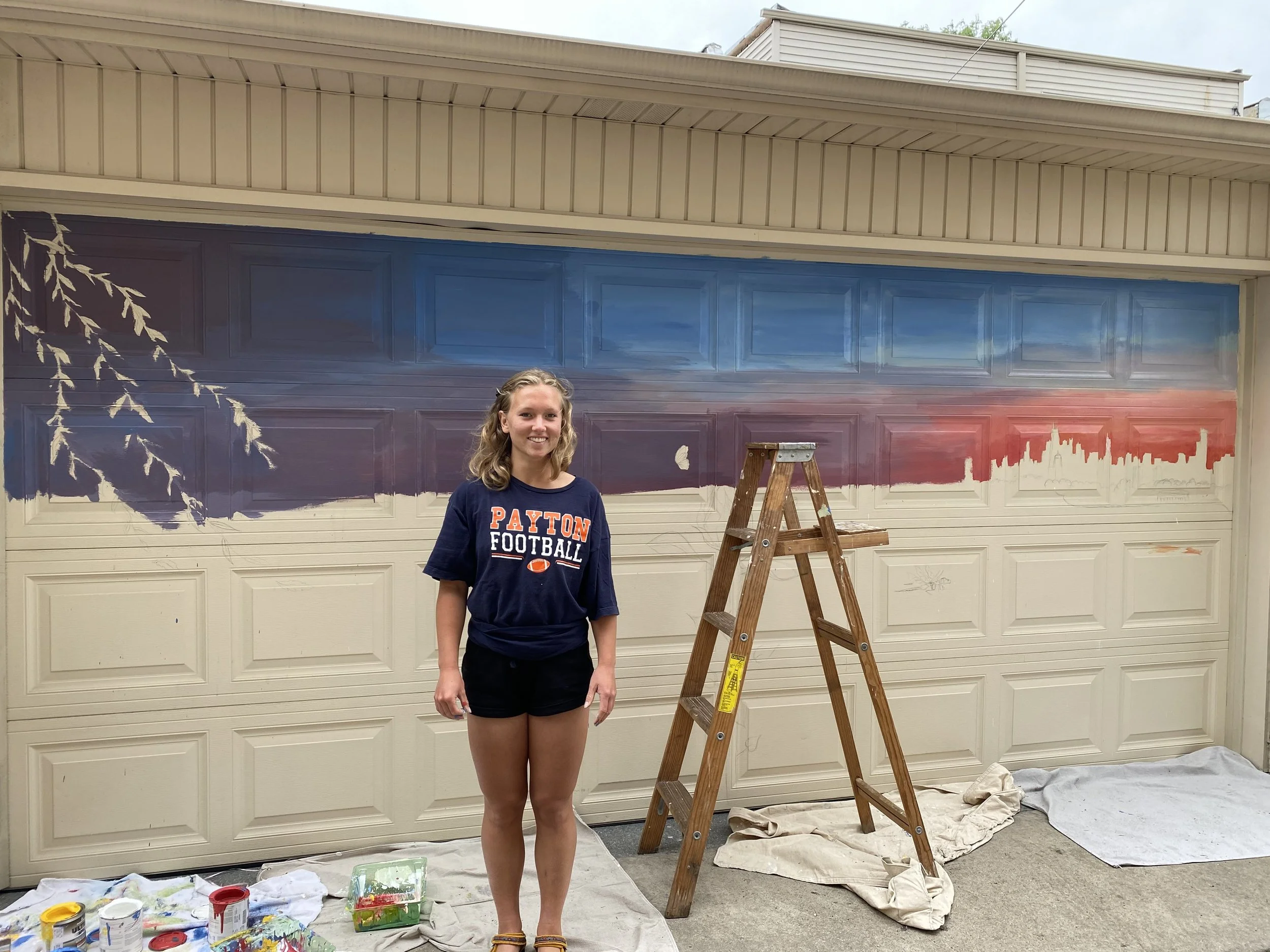 Young woman wearing a navy blue Payton Football T-shirt stands in front of a garage door with a partially painted mural in progress, showing a skyline and trees. An open wooden ladder and painting supplies are on the ground.