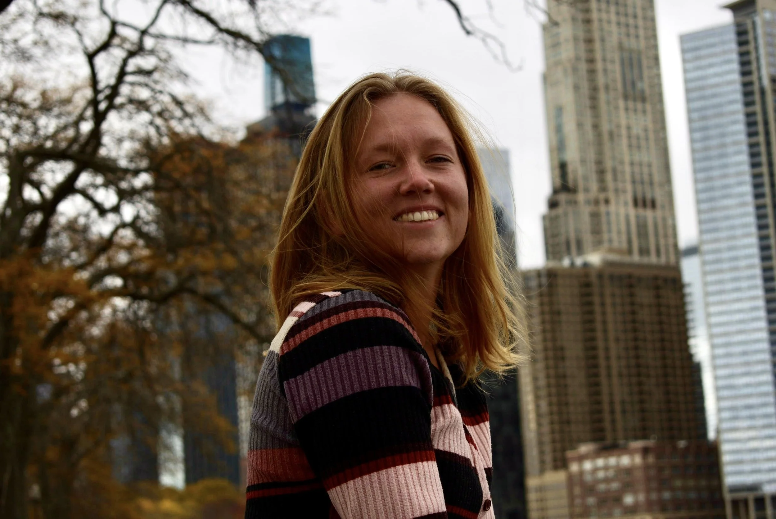 A woman with long blonde hair smiling outdoors with a city skyline and trees in the background.