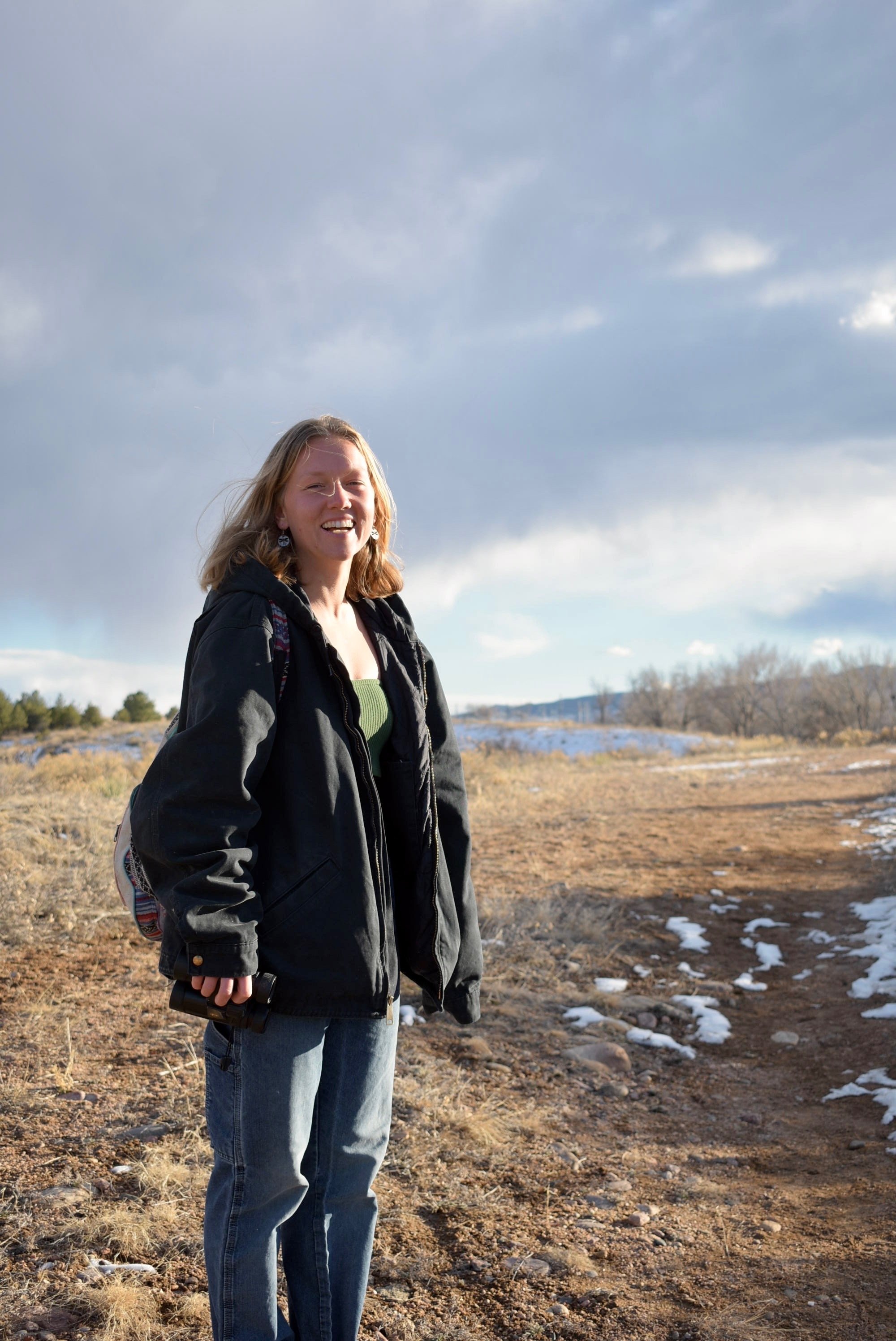 A young woman standing outdoors on a dirt path in a landscape with patches of snow, wearing a black jacket, green top, and jeans, smiling at the camera with cloudy skies overhead.
