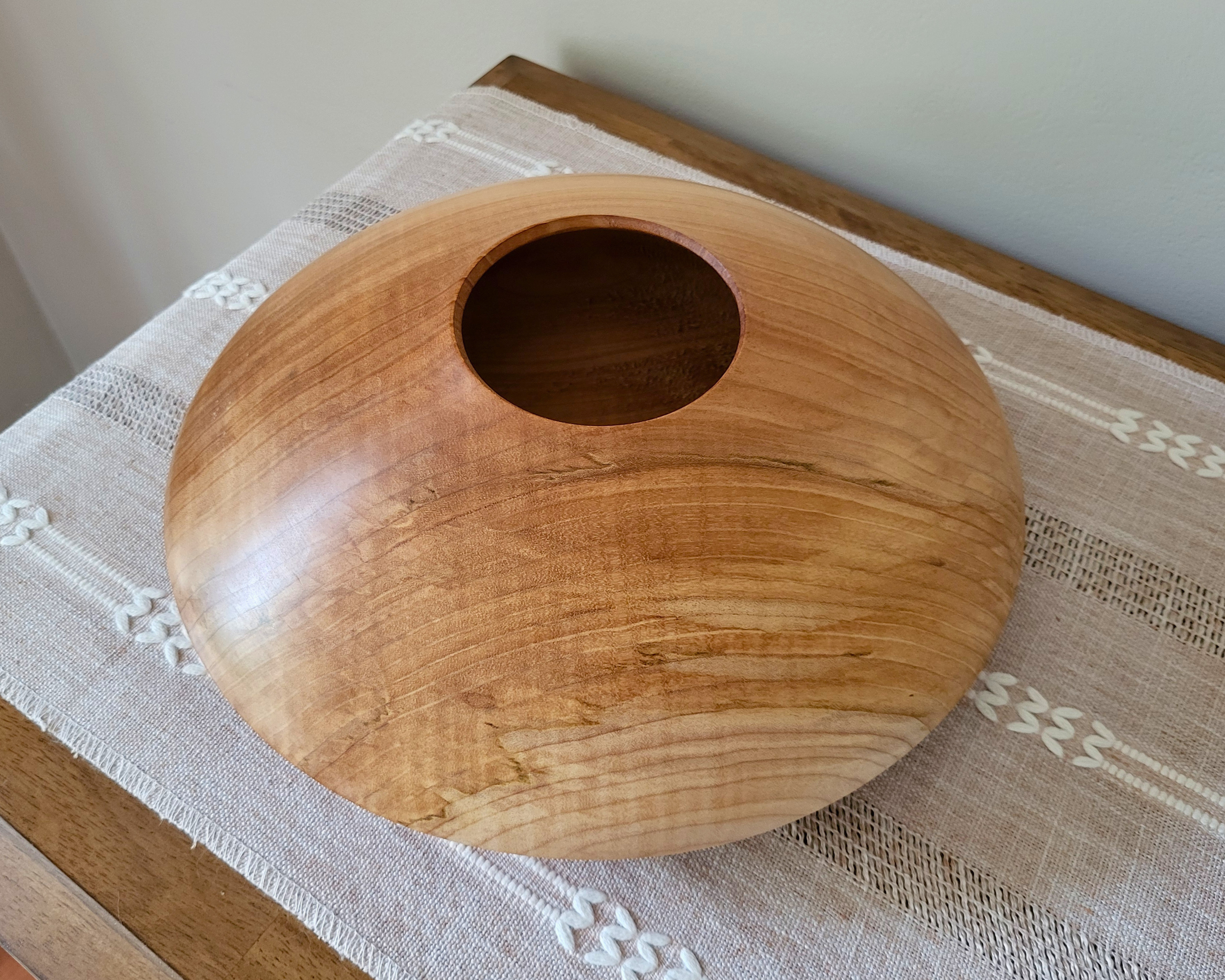 Angled overhead view of a curly maple closed-form hollow vessel by Treverk Woodturning displayed on a woven table runner, with tight curly figure and the small centered opening clearly visible. One of a kind sculptural wood art.