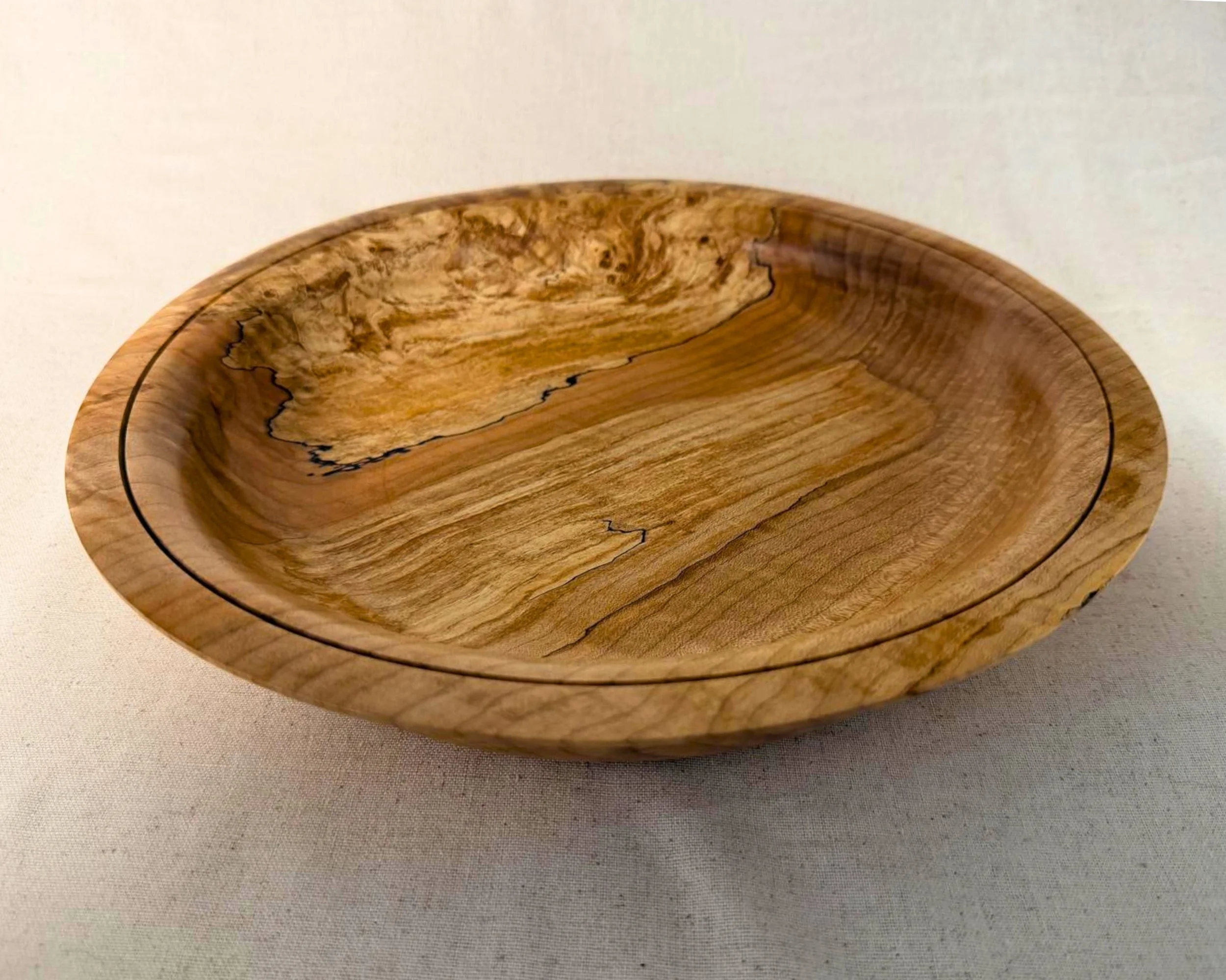 A round wooden bowl with natural grain and a slightly uneven edge, sitting on a neutral fabric background.