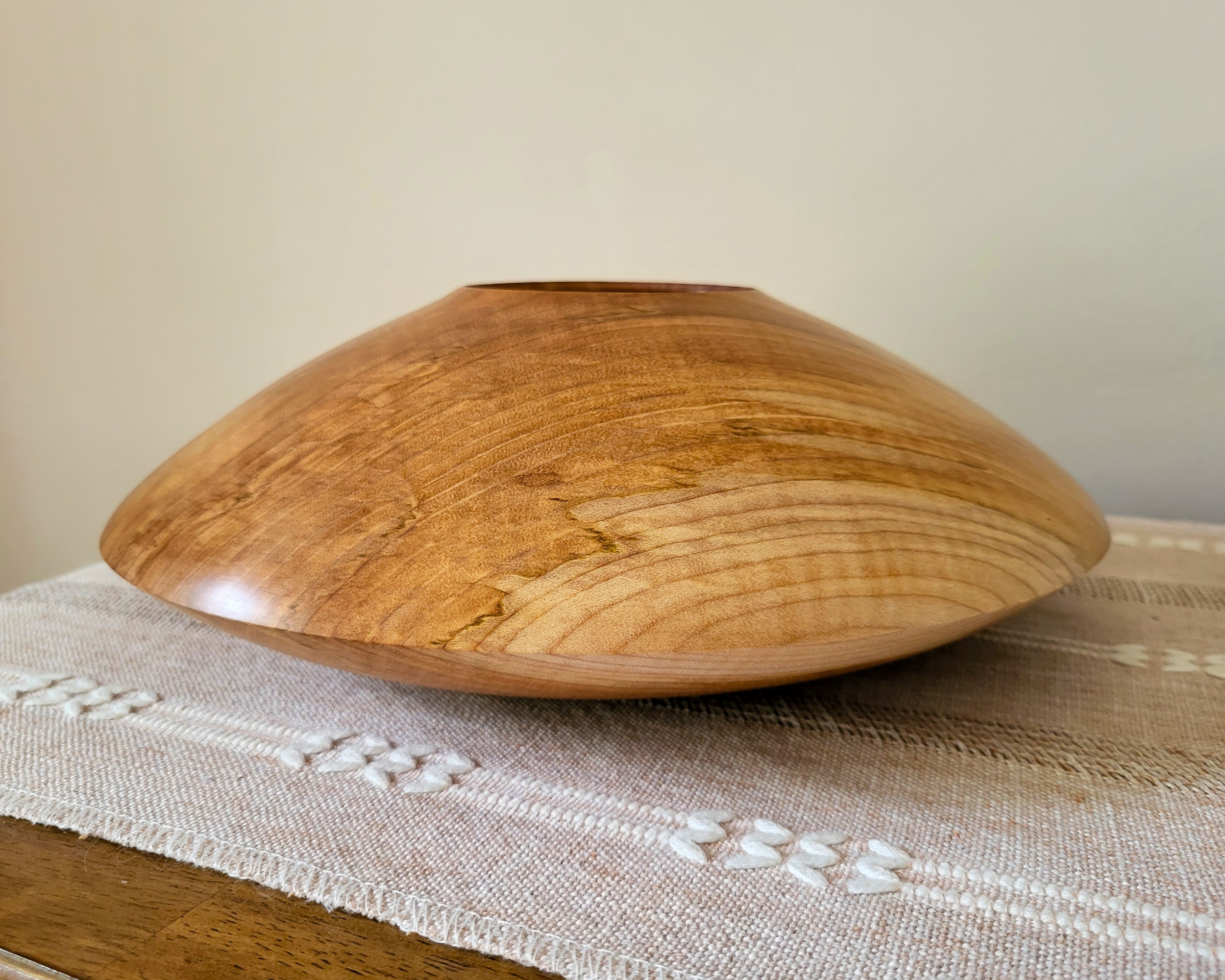 Low, wide curly maple closed-form hollow vessel by Treverk Woodturning sitting on a woven linen table runner, showing the lentil-shaped profile in soft matte satin finish. Sculptural wood centerpiece for a coffee table or console table.