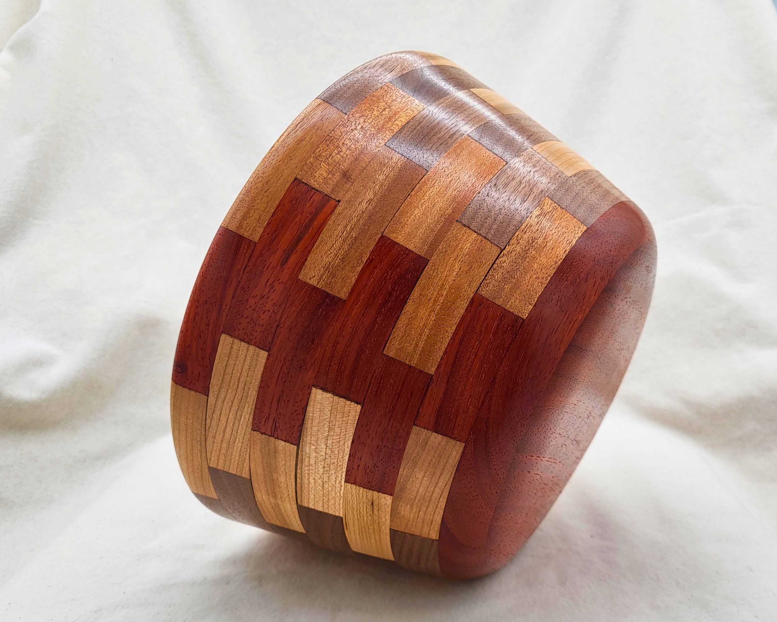 Side angle view of a hand-turned segmented wood bowl by Treverk Woodturning, tilted to show both the curved base and the basket-weave wall pattern in padauk, maple, mahogany, and walnut. Sculptural decorative wood bowl.