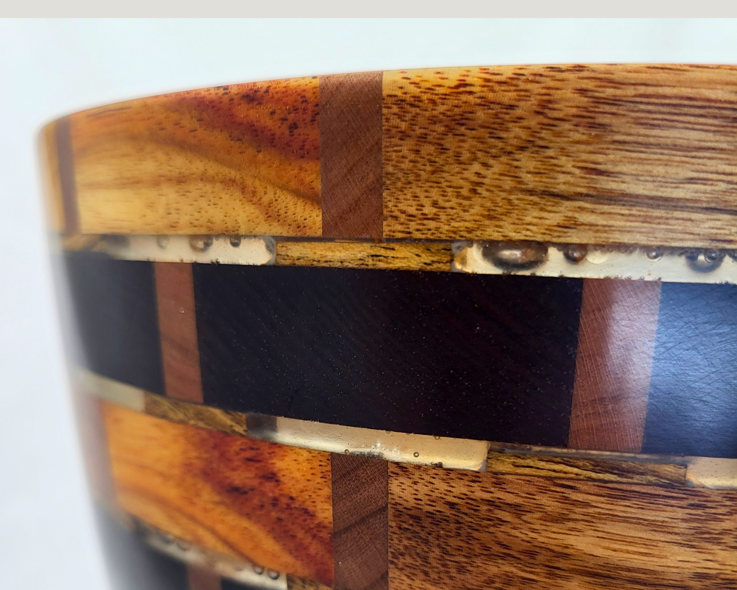 Close-up of the rim and upper wall on a hand-turned segmented vessel by Treverk Woodturning, showing canarywood blocks with rust and gold streaks, narrow cherry pillars, charcoal thermal ash bands, and clear resin between every joint.