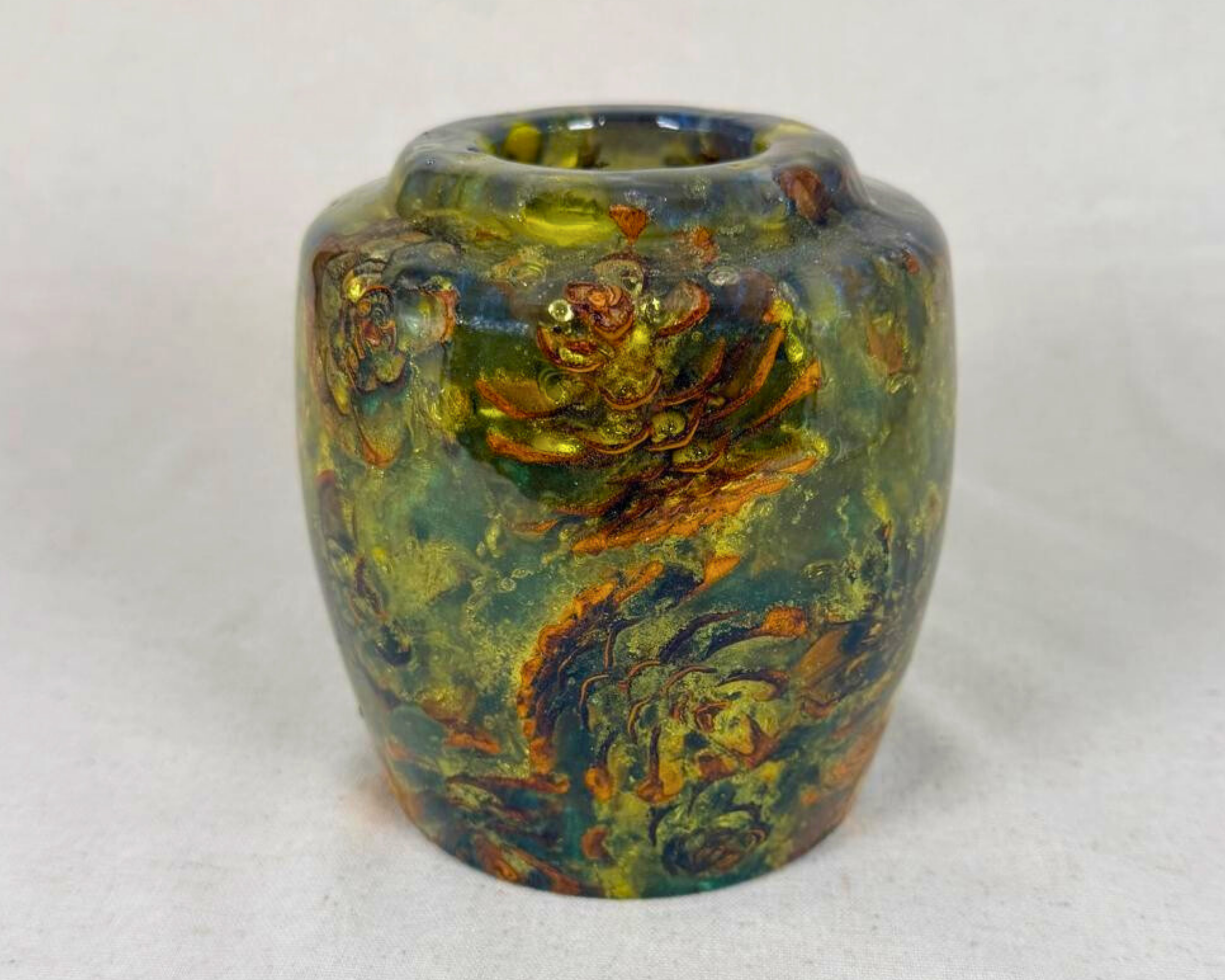 Pine cone vessel in swirling green, teal, blue, and gold epoxy, photographed front-facing to show its luminous, underwater effect and clustered natural inclusions.