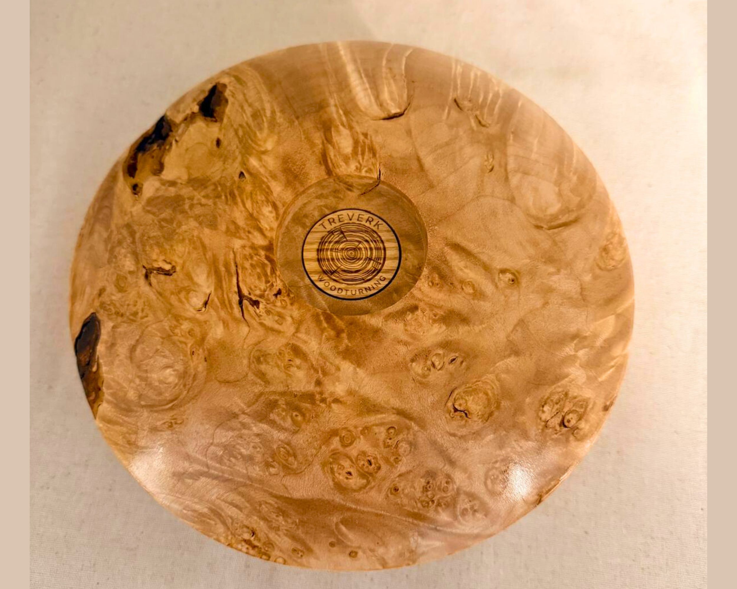 Bottom view of a hand-turned maple burl vessel showing dense burl figure, scattered natural inclusions, and a centered Treverk Woodturning mark.