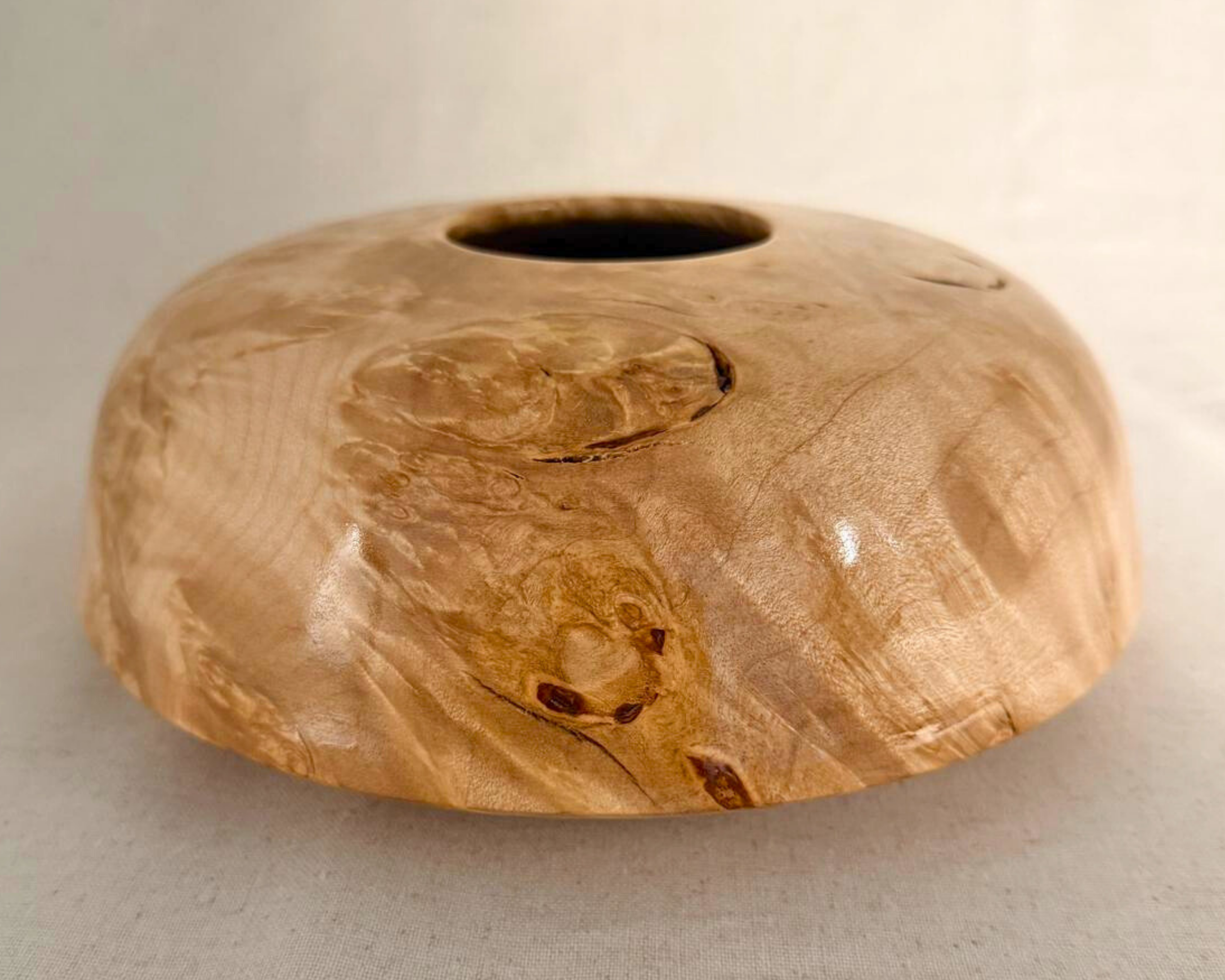 Side view of a low, wide maple burl vessel with a small opening and a dramatic dark natural void cutting across the body, creating a floating visual effect.