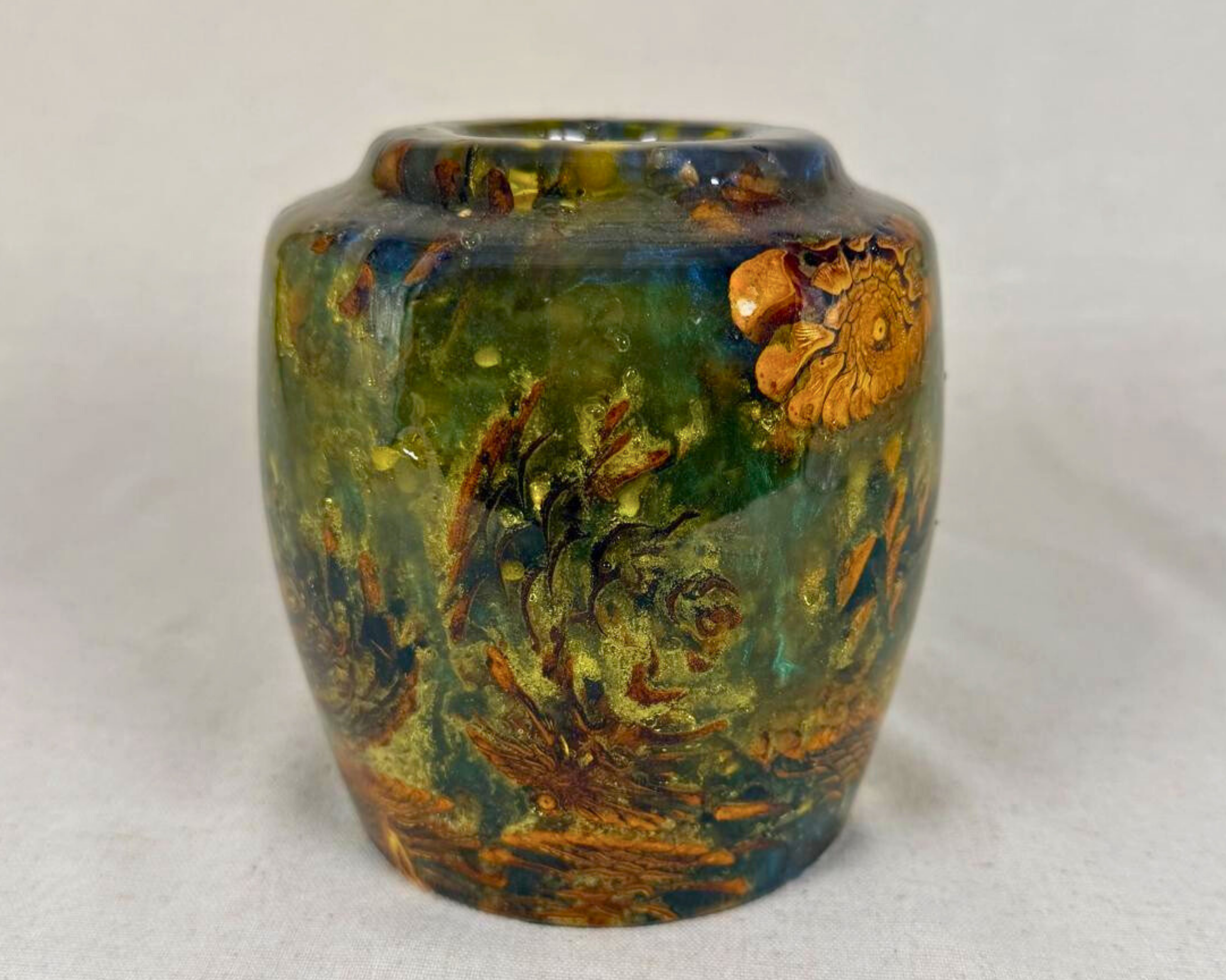 Hand-turned pine cone and epoxy vessel in deep olive green, teal, blue, and amber tones, shown front-facing with an underwater-like glow and a rounded shoulder.