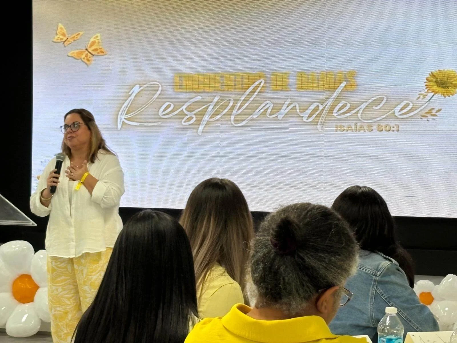 A woman with glasses and a white shirt speaking into a microphone in front of a large screen that says 'Encourage the Daughters' with a Bible verse reference. Audience members are sitting and listening, with white and orange balloons on the table.