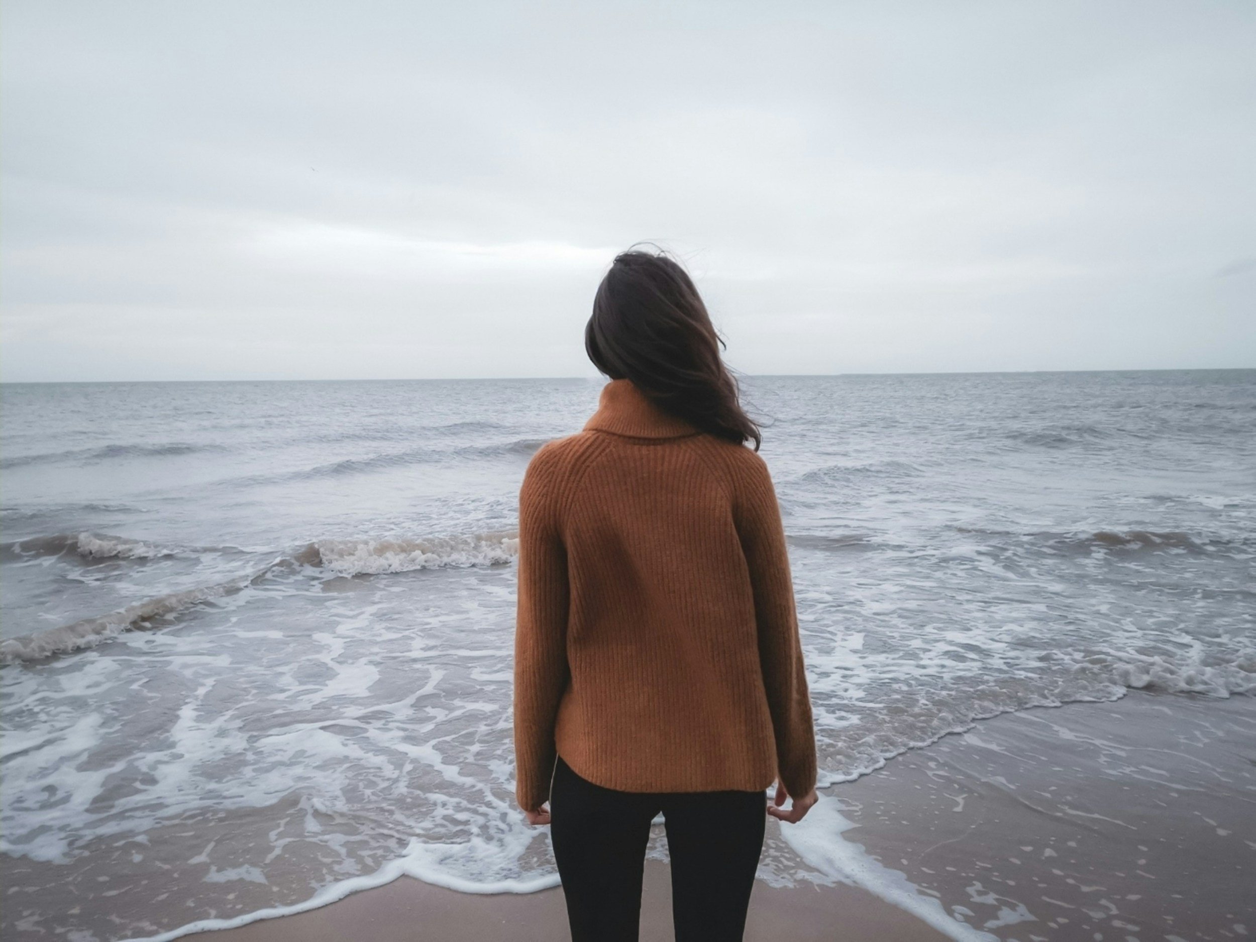 Woman standing facing the ocean wearing long sleeve blouse and black pants