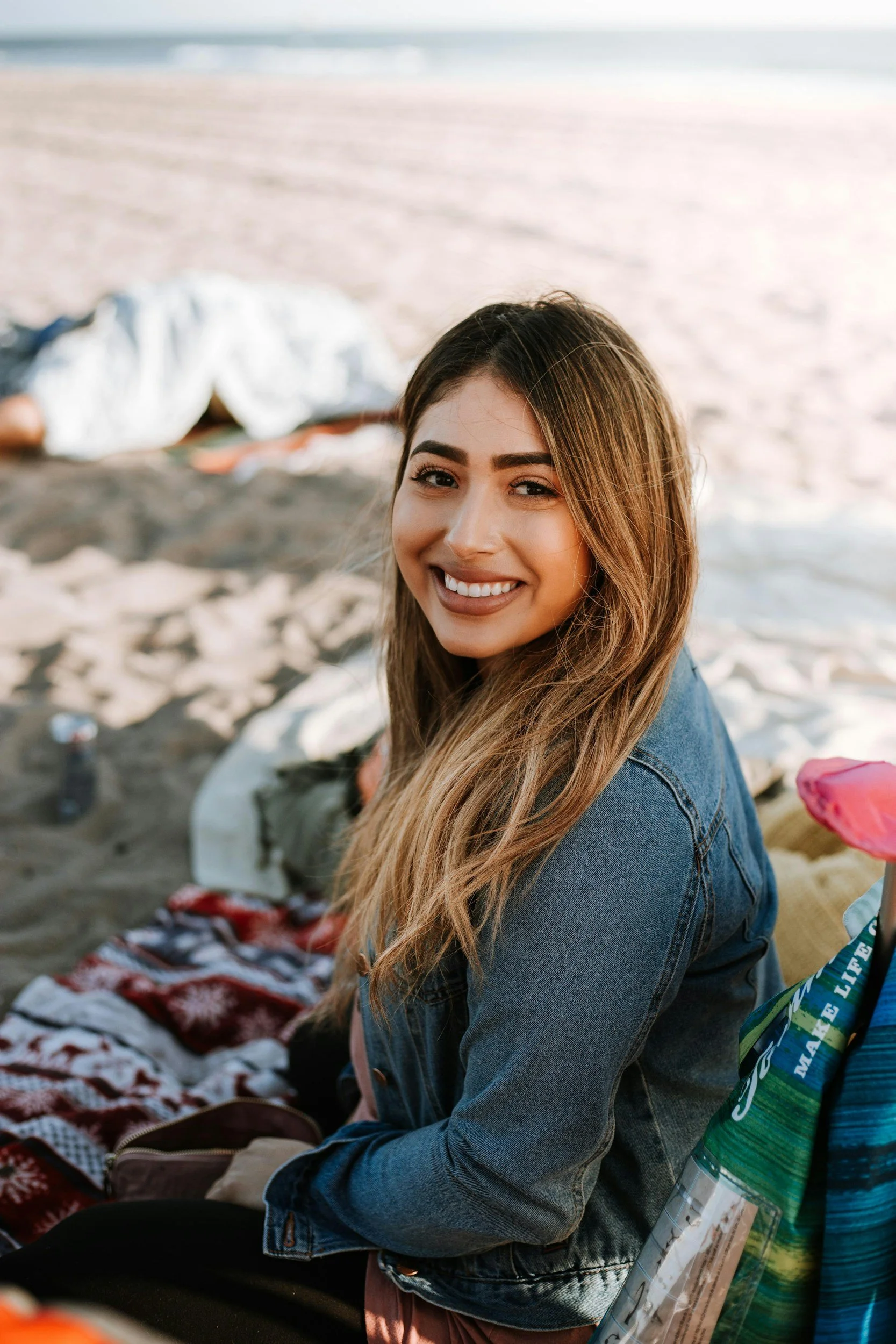 Young woman smiling at the beach during sunset, sitting on a blanket with a person lying in the background, surrounded by beach items.