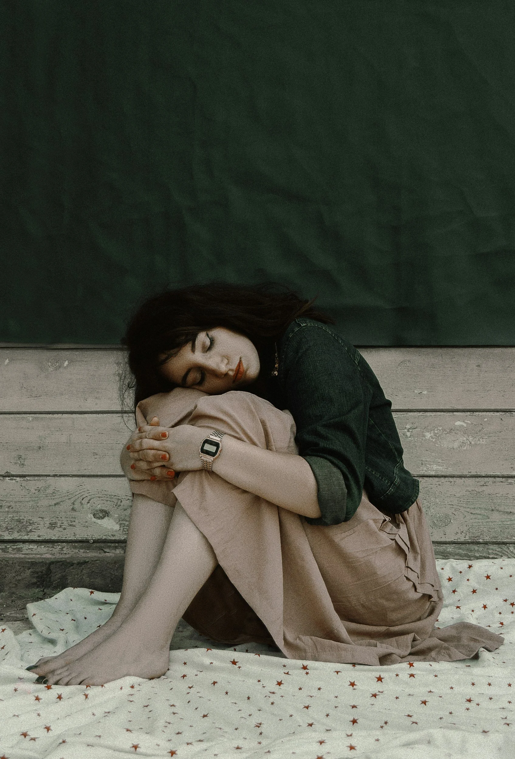 A woman with dark hair and red lipstick, wearing a dark green denim jacket and beige skirt, sitting on the floor with knees bent, hugging her shins, resting her head on her knees with her eyes closed.